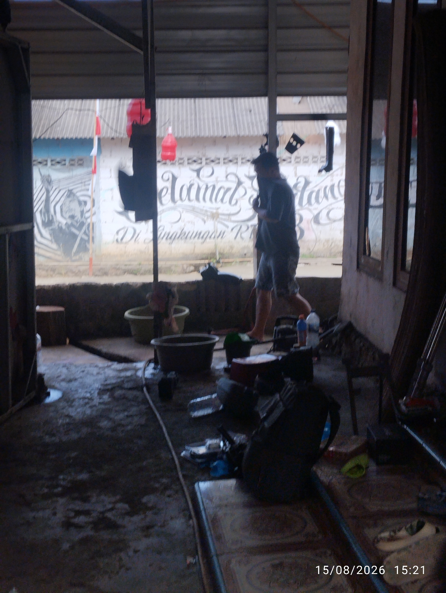 The image captures a dimly lit, cluttered outdoor area, possibly a workshop or storage space, with a concrete floor that appears wet and dirty. In the background, a mural on a wall depicts a zebra and text that is partially obscured by shadow, but appears to read "Selamat Datang Di Lingkungan RT" which translates to "Welcome to the RT Environment." Red plastic bottles are hanging from a structure, and two Indonesian flags are visible.
A person, appearing to be male, is walking from left to right in the mid-ground, wearing a dark t-shirt and shorts, and is barefoot. The environment is filled with various objects including buckets, tools, hoses, bags, and what appear to be discarded items. The overall impression is of a utilitarian or work-in-progress space. The timestamp at the bottom right indicates the image was taken on August 15, 2026, at 15:21.
