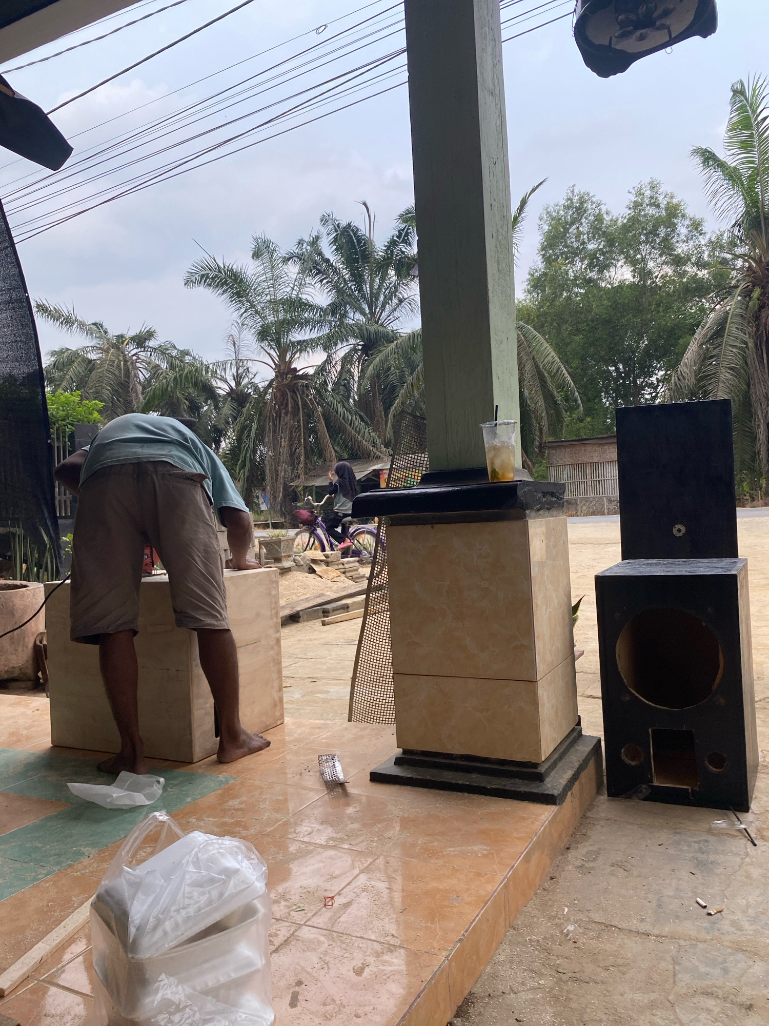 In Mojo Pahit, Indonesia, a man, wearing a light blue t-shirt and brown shorts, is seen from behind working on a large, unfinished wooden box. He appears to be sanding or attaching something to its surface with a tool, his bare feet placed on the tiled ground. Beside him, a black speaker cabinet stands upright, and another larger, empty speaker enclosure rests on its side. In the background, palm trees line the street, with a building and a person on a purple bicycle visible further down. Overhead, power lines crisscross the cloudy sky. A discarded plastic cup with a beverage sits on a tiled pedestal. The scene suggests an outdoor workshop or construction area.