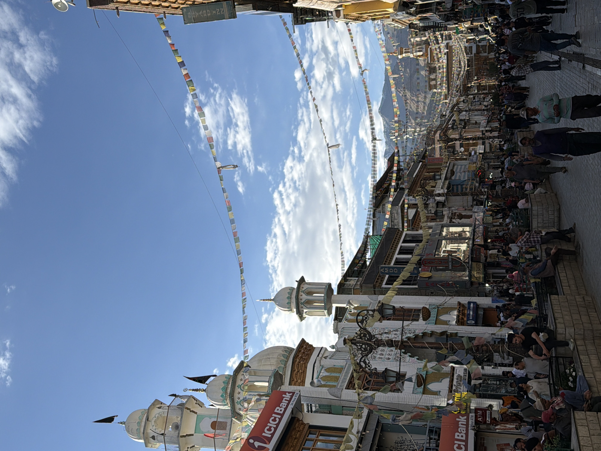 The image depicts a busy street scene in the city of Leh, India. The street is lined with buildings, many of which appear to be shops and businesses, with signs such as "ICICI Bank" and "GOLDEN BAKERY" visible. Prayer flags are strung across the street, and two prominent minarets, likely part of a mosque, stand on the left side of the image. Mountains are visible in the background. A crowd of people are walking and standing on the street, some appearing to be shoppers or tourists, while others are seated on a low wall. The sky is blue with scattered clouds, and the sun appears to be shining.