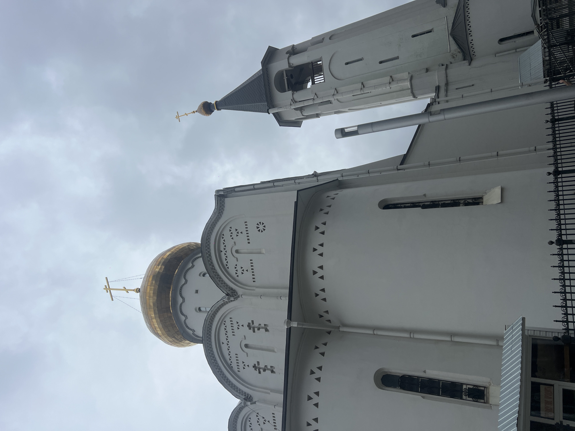 The image shows the architectural details of a Russian Orthodox church in Moscow, Russia. The angle of the shot is looking upwards towards the white facade of the building. The church features a golden dome with a prominent cross at its apex. The exterior walls are adorned with decorative patterns and religious symbols. Adjacent to the church structure is a tall bell tower, also painted white, with open arches towards the top. The sky is overcast. A metal fence is partially visible, hinting at the surroundings of the church.