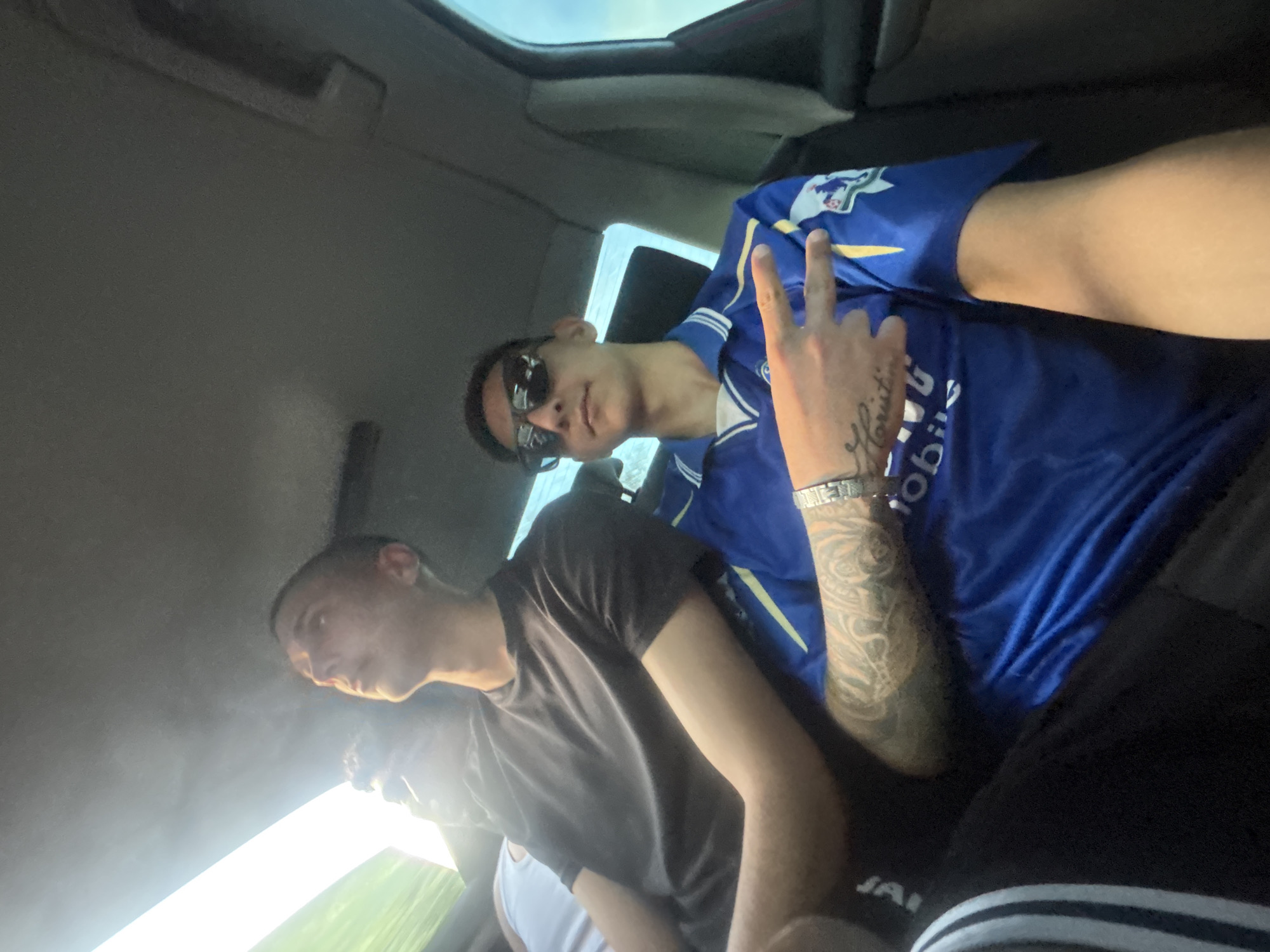 The image shows three young men inside a vehicle. One man is wearing a blue jersey with a visible tattoo on his arm and sunglasses, holding up a peace sign. Another man is wearing a dark gray shirt. A third man is visible partially, wearing a white shirt. The environment appears to be the interior of a car, with the sun shining through the windows. The location in the city is Unknown, Unknown.