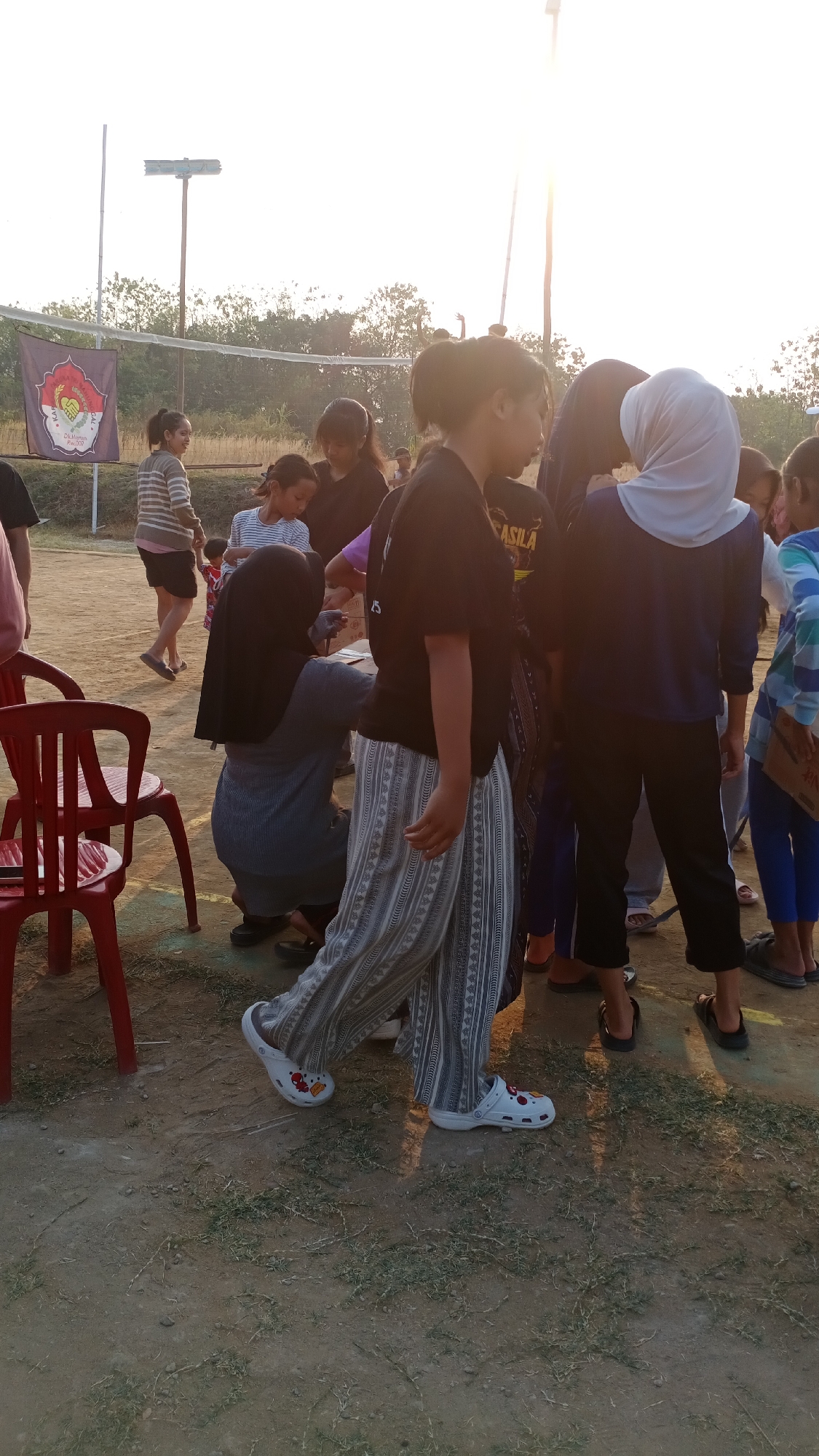 The image depicts a group of people, likely young individuals, gathered outdoors in what appears to be a casual setting. The ground is dirt with patches of dry grass, suggesting an unpaved area such as a field or a yard. In the background, trees are visible, and a volleyball net is strung across, indicating a possible recreational space. A banner with an emblem and text is also present. The lighting suggests it is either early morning or late afternoon, with the sun casting a warm glow.
Several individuals are the focus of the image. In the foreground, a person wearing a black t-shirt with "ASILA" written on it and patterned grey and white pants is walking, showing off white clogs with cartoon characters. To the left, a person in a grey top and black hijab is kneeling, seemingly interacting with a paper or surface. Other individuals are standing around them, some wearing casual clothing, including a striped sweater and shorts, and others in athletic wear. One person is wearing a light blue headscarf. The overall atmosphere seems relaxed and informal. While the prompt mentions the city of Mertan, Indonesia, this specific location cannot be definitively identified from the visual information alone. However, the scene is consistent with outdoor public spaces in Indonesia.