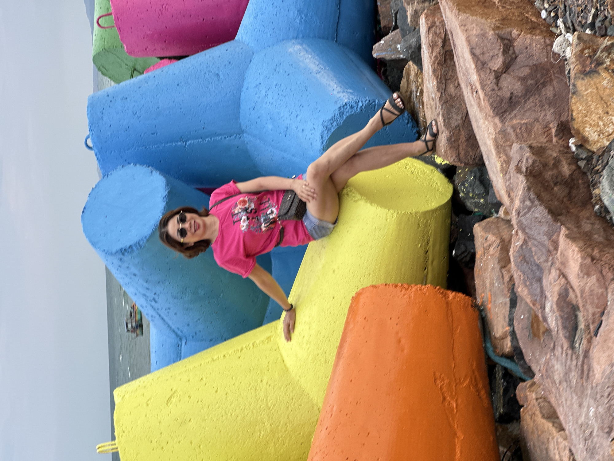This media file captures a vibrant outdoor scene in Phu Thuy Ward, Vietnam, centered on a woman posing amidst large, colorful concrete structures. The image is oriented vertically, showing the woman lying on her back across bright blue and yellow blocks. She is smiling directly at the viewer, wearing a pink t-shirt with a graphic design, denim shorts, black sandals, and sunglasses. Her left hand rests on a yellow block, while her legs are casually extended and bent, showcasing her playful pose.
The setting is a coastal area, likely a breakwater or pier, where numerous large, geometrically shaped concrete blocks are stacked. These structures, often referred to as tetrapods or wave-breakers, come in various bold colors, including blue, yellow, pink, green, and orange, creating a striking contrast with the natural environment. To the right of the frame, large, reddish-brown rocks are visible, forming part of the shoreline or a natural revetment. In the background, a body of water (likely the sea or ocean) stretches under a pale, overcast sky, with a small, distant boat visible on the horizon. The overall mood conveyed by the woman's smile and casual pose is one of relaxation and enjoyment. No clearly legible text is visible on any objects or in the background.