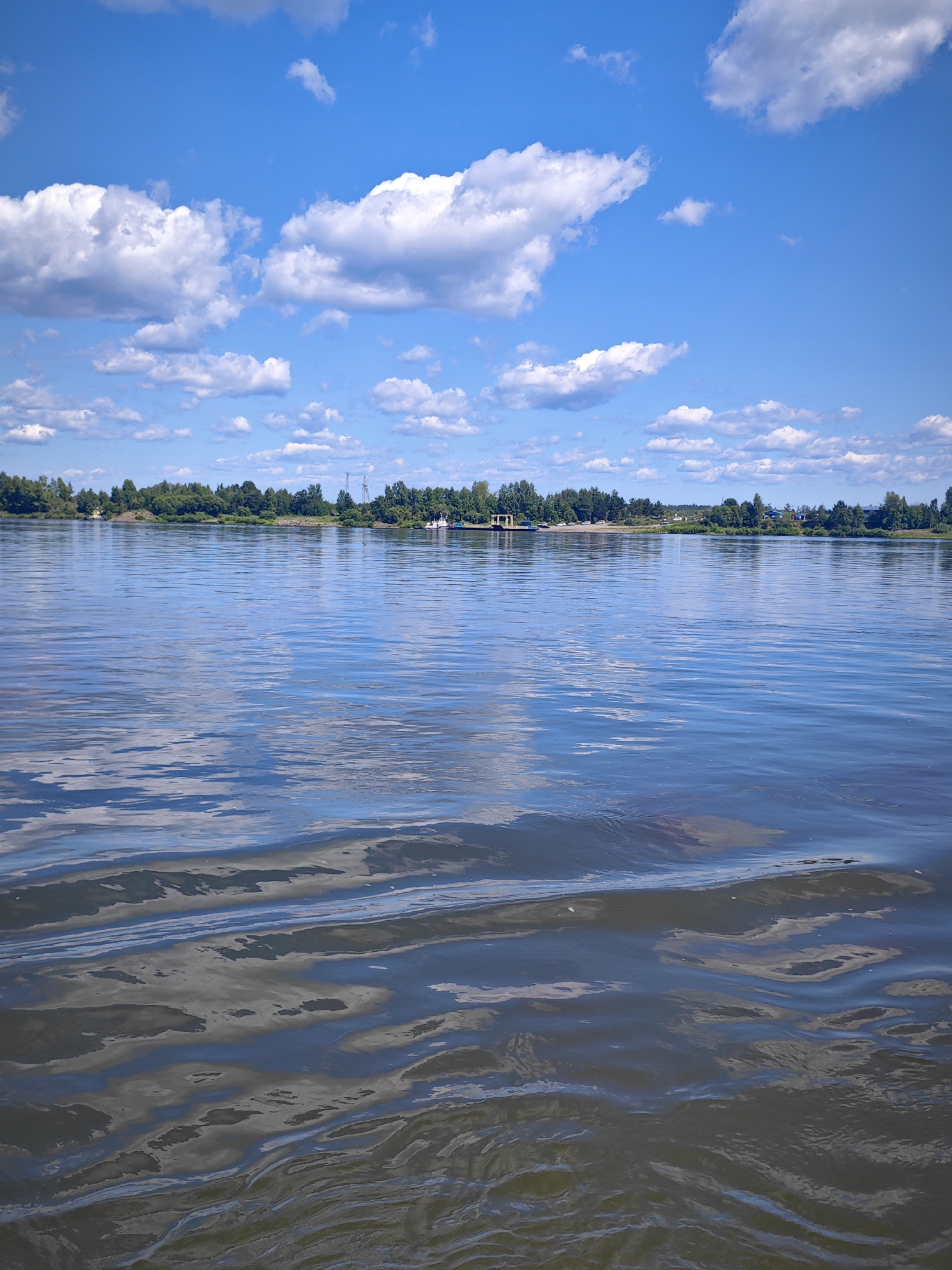 The image shows a wide body of water, likely a river, on a clear day with scattered white clouds in a bright blue sky. The water has ripples and reflections of the sky and the distant shoreline, which is lined with a dense forest of green trees. In the distance, a few structures and possibly a small settlement can be seen along the tree line. There are no people or specific actions depicted. The scene conveys a sense of natural landscape and tranquil weather. The location is identified as Krutoi Log, Russia.