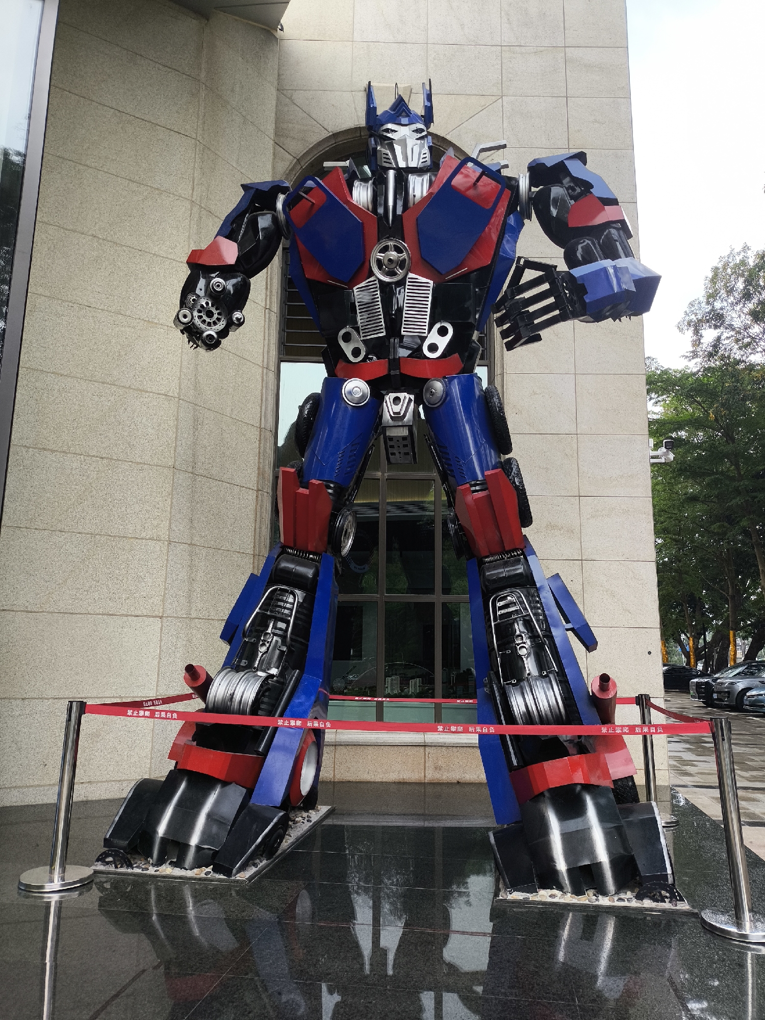 The image shows a large, colorful sculpture of Optimus Prime, a character from the Transformers franchise. The sculpture is predominantly blue, red, black and silver, with a detailed metallic finish. It stands in front of a modern building with a light beige stone facade. A red safety barrier with white Chinese characters on it surrounds the base of the sculpture. The ground around the sculpture is a dark, reflective surface, possibly marble.
In the background, beyond the sculpture, there are green trees and a glimpse of parked cars, suggesting an urban environment. The scene is likely located in Longgang District, China. The image seems to have been taken during the day, with overcast lighting. There are no people visible in the image. The sculpture dominates the scene, conveying a sense of scale and artistic display.