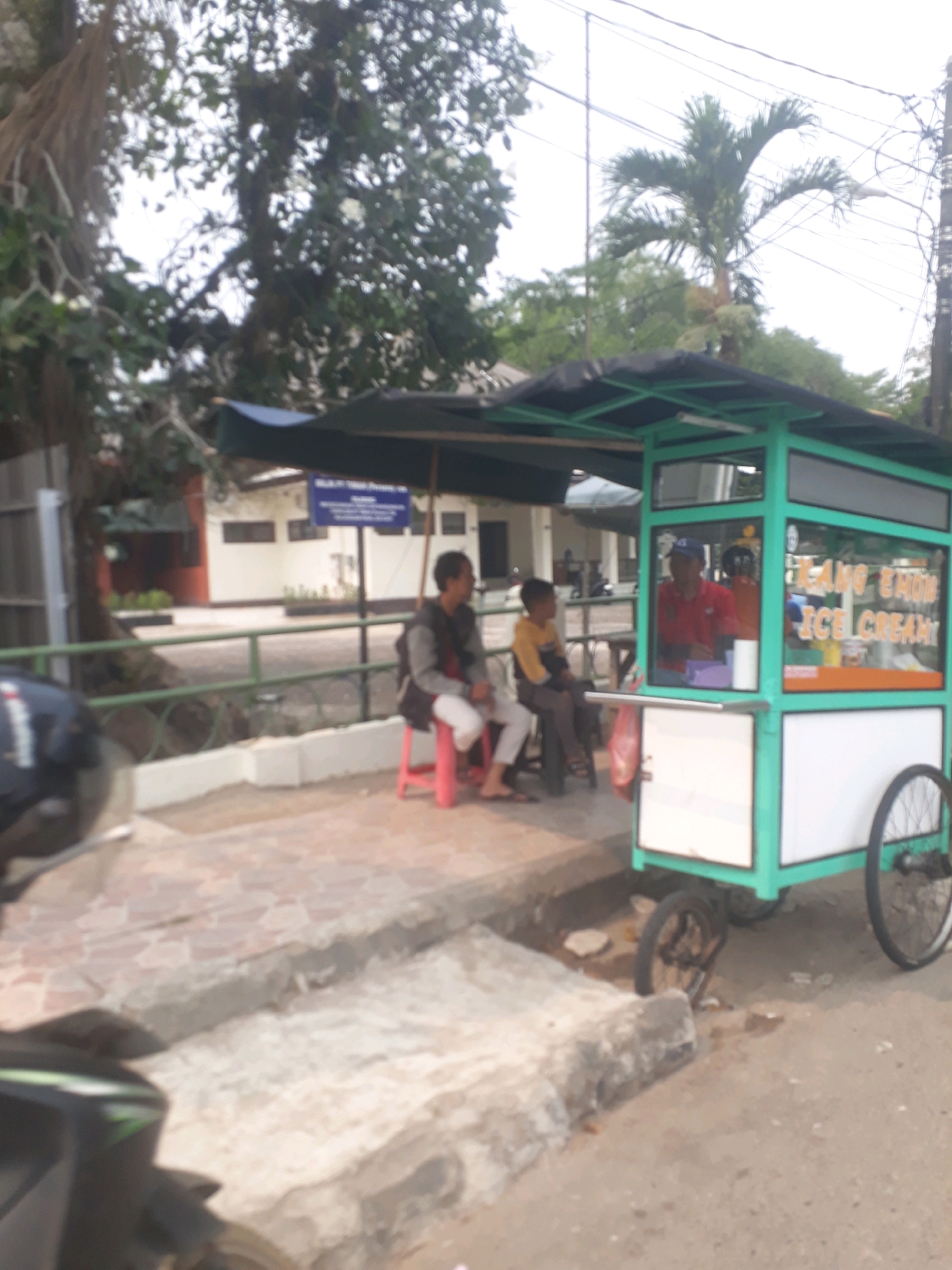 The image captures a scene on a street in Pangkalpinang, Indonesia, in front of a building with light-colored walls and dark-framed windows. A green ice cream cart with a black awning is the central object. The cart displays "MAKMUR JAYA ICE CREAM" in yellow lettering on its side. A man in a red shirt and blue cap is visible inside the cart, presumably serving. Two other individuals are seated on red stools outside the cart. To the left of the ice cream cart, a portion of a black motorcycle is visible, and further left, a large tree with exposed roots dominates the background. The foreground shows a paved sidewalk with red bricks and a gray concrete curb. The overall setting appears to be a commercial or public area.