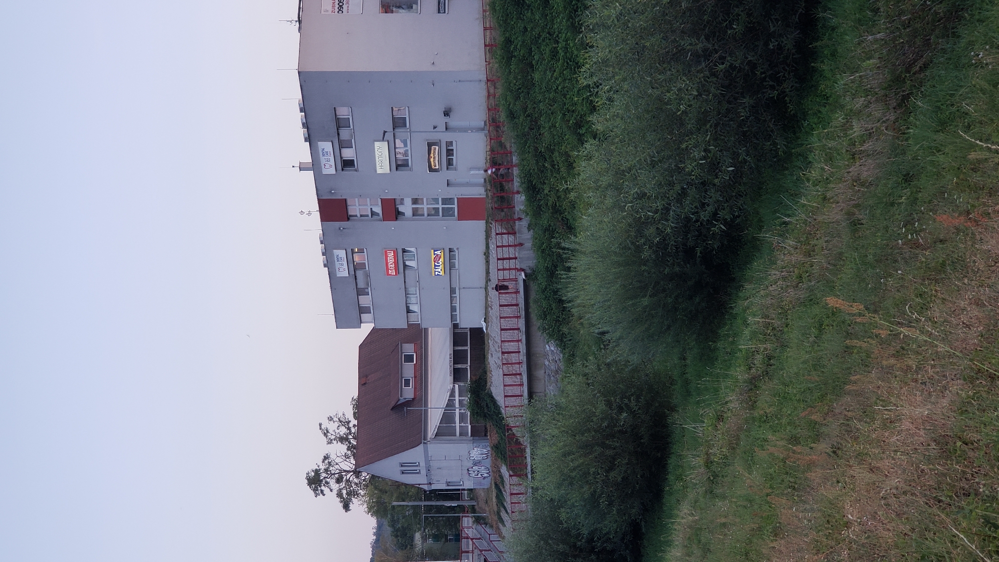 The image shows a modern, multi-story building with large windows and signage, located in the city of Prešov, Slovakia. The building is grey with red accents and has various signs displaying company names such as "DENTAL", "GENERALI", "HARMONY", and "ZÁLOŽŇA". To the left of this building is a smaller, older structure with a brown roof and graffiti on its exterior wall. In the foreground, there is lush green foliage and grass bordering a body of water, likely a river or canal. A red pedestrian bridge with railings spans across the water, connecting the two buildings and the surrounding area. Two individuals are visible on the bridge, one walking and the other running. The sky is a pale gradient of blue and white, indicating either early morning or late evening.