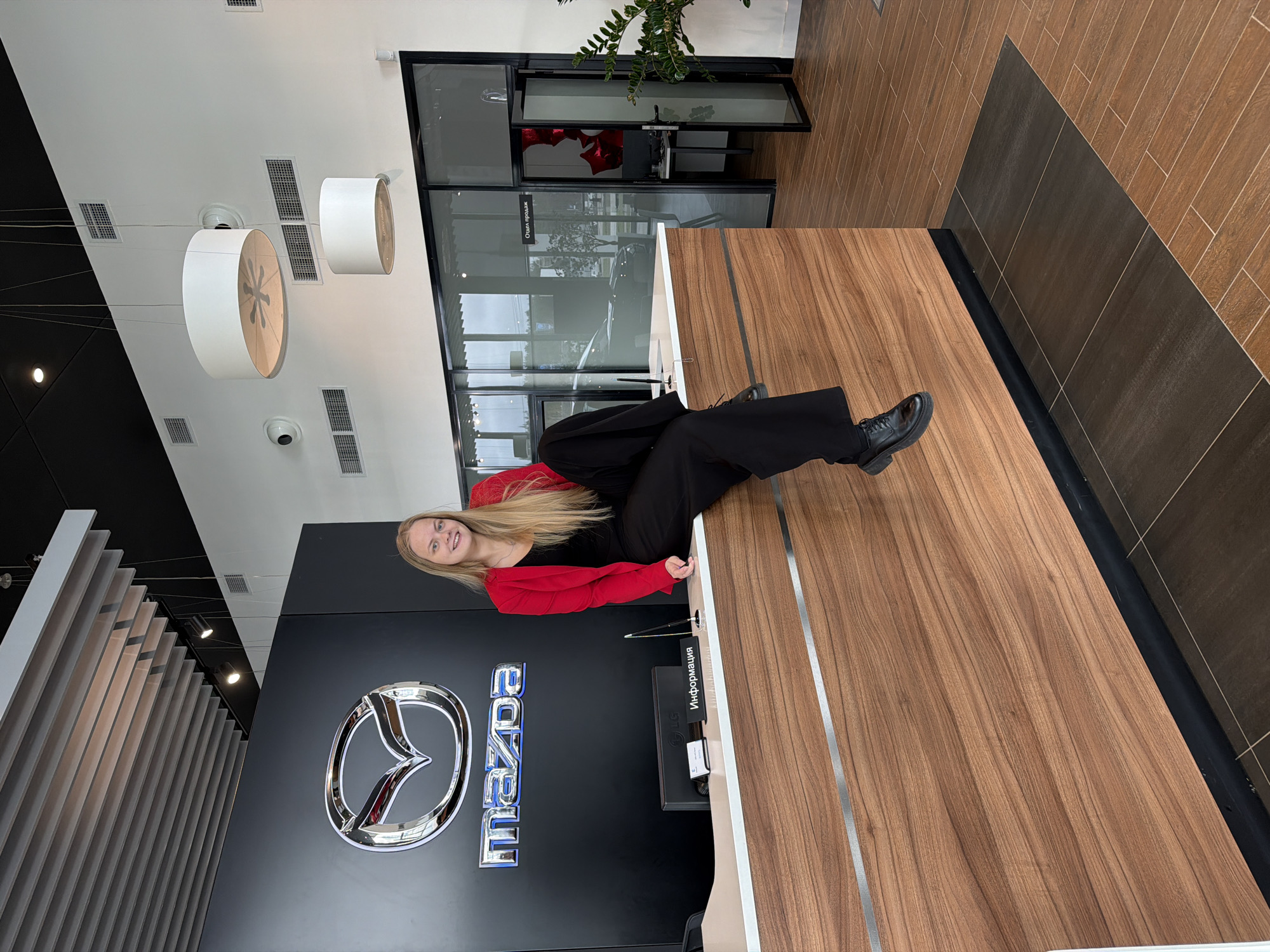 A young woman stands smiling inside a Mazda dealership in Izhevsk, Russia. She is positioned behind a wooden counter, wearing a red blazer and black pants. Behind her is a black wall with the silver Mazda logo and lettering. The dealership has modern lighting fixtures hanging from the ceiling. A glass-enclosed room is visible in the background, and the floor is tiled in a wood-grain pattern with a darker strip along the edge.