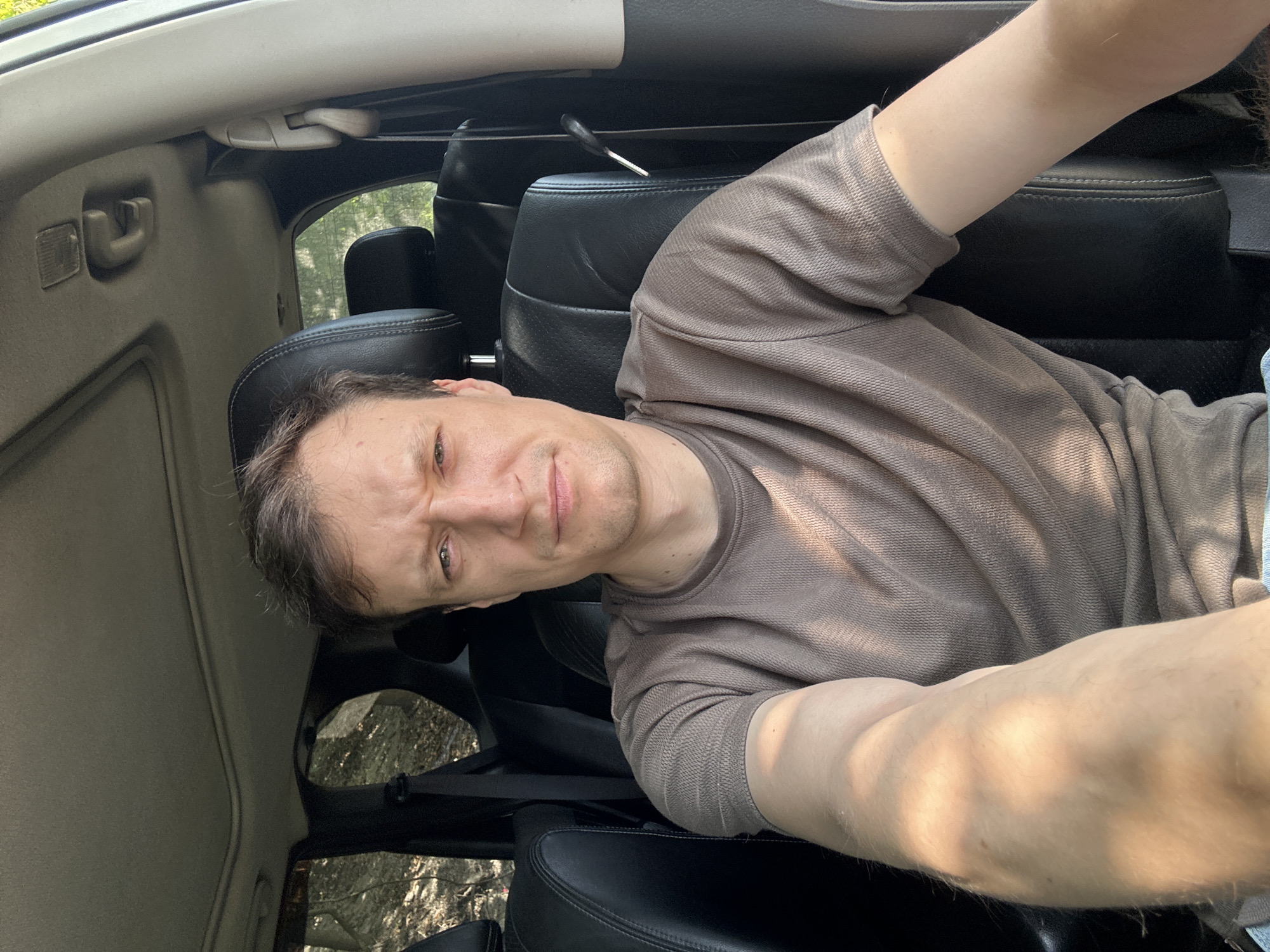 A medium shot shows a man sitting in the driver's seat of a car, presumably in the city of Makiv, Ukraine. He is looking directly at the camera with a furrowed brow and a slightly pursed mouth. His left arm is extended toward the camera, holding it for a selfie. The man is wearing a plain, taupe-colored t-shirt. The interior of the car is visible, with black leather seats and a sunroof overhead. Through the windows, trees and foliage can be seen, suggesting the car is parked outdoors, possibly in a wooded or rural area. The lighting indicates it is daytime, with sunlight casting shadows on the man's arm and face.