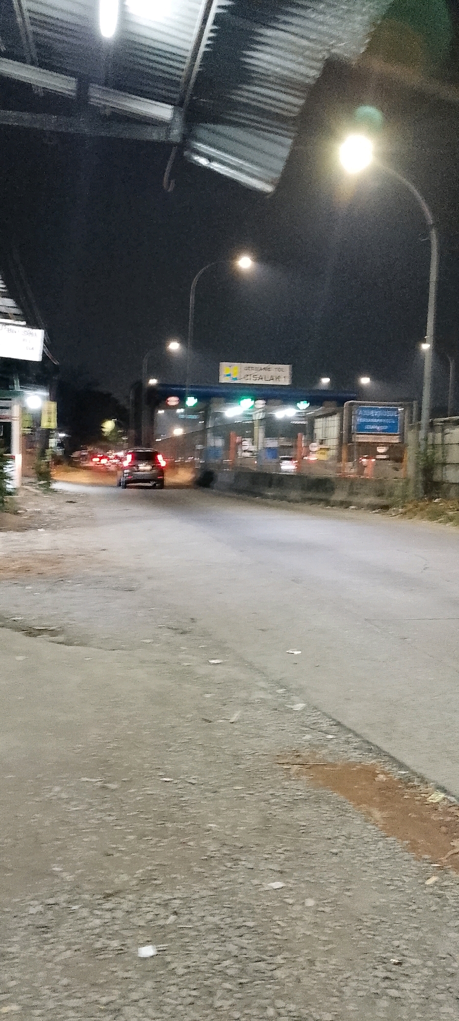 This is a night-time photograph taken at the Cisalak 1 Toll Gate in Depok, Indonesia. The image shows a toll plaza with several lanes, some of which are open with green lights indicating they are functional. A white sign with blue lettering that reads "GERBANG TOL CISALAK 1" is visible above the lanes. There are streetlights illuminating the area, and the sky is dark. A few cars are visible on the road, with one car in the foreground appearing to be a dark-colored SUV. The road surface is asphalt, and there is some debris scattered on the side of the road. The overall scene depicts a typical night-time view of a toll gate on a highway.