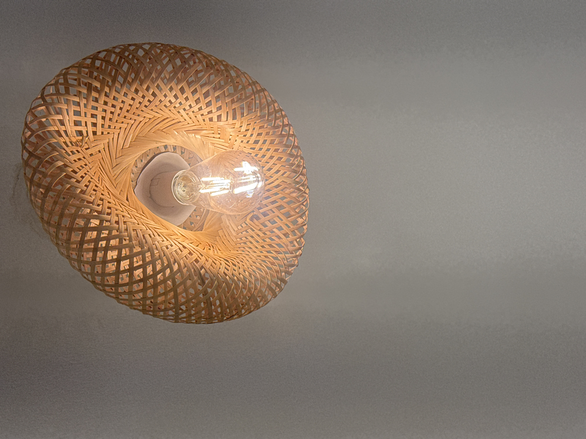 A ceiling light fixture with a woven bamboo shade hangs from a neutral-colored ceiling. The light fixture has a clear Edison-style bulb. The background is a plain, textured wall. The image does not provide any information about the city of Abuja, Nigeria.