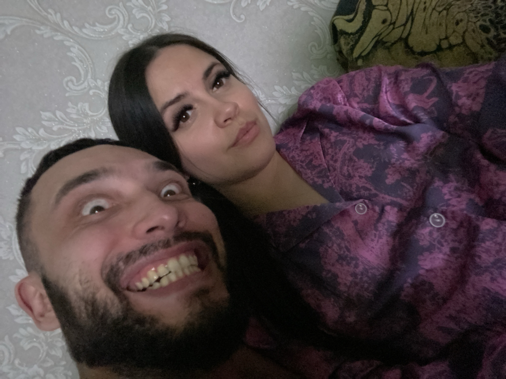 A man and a woman are positioned closely together, likely lying down. The man is in the foreground and is making an exaggerated expression with his eyes wide and mouth open, exposing his teeth. He has dark hair and a beard. The woman is behind him and appears to be looking at the camera with a more subdued expression. She has dark hair and makeup.
The background includes a patterned wall. The woman is wearing a patterned garment with buttons. There are no identifying elements to pinpoint the location as Chaikovsky, Russia or any specific city.