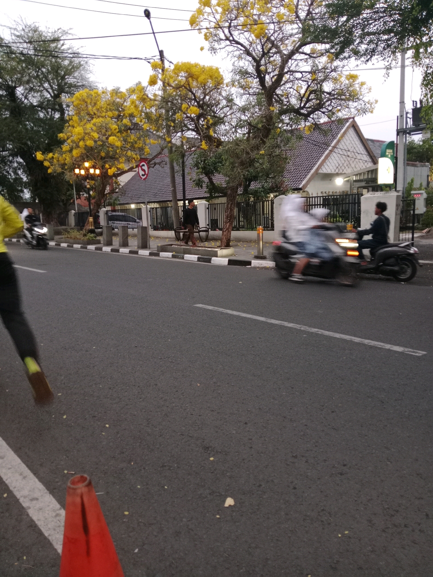 This is a street-level, eye-level, medium shot of a street in Sawah Gede, Indonesia, during the day. A person in a yellow top and dark pants is running on the left side of the road. In the middle of the road, two motorcycles are in motion. The first motorcycle, on the left, is carrying a person wearing a light-colored hoodie and dark pants. The second motorcycle, on the right, is carrying a person in a dark top and pants. The road is paved and gray, with white lines marking lanes. On the right side of the road, there is a sidewalk with a black and white striped border. Behind the sidewalk, there is a house with a white facade and a gray tiled roof. Yellow flowers are blooming on trees lining the street. A traffic light and street lights are visible, along with a street sign with a red circle and an 'S' inside, indicating a restriction. An orange traffic cone is in the foreground on the left.
