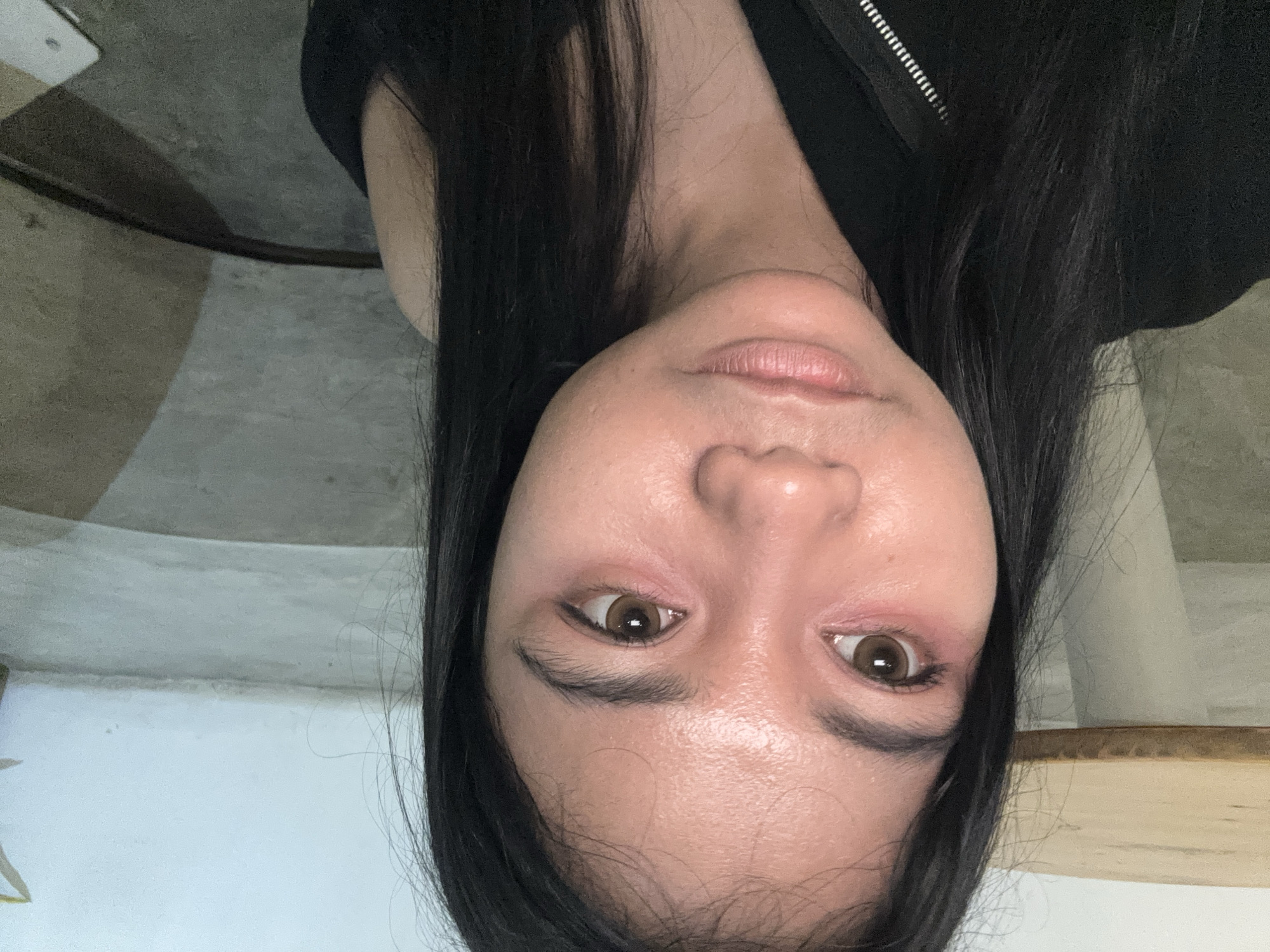 A close-up, inverted perspective of a woman's face, revealing light brown eyes with visible eyeliner, eyebrows, and pink eyeshadow on the eyelids. Her skin shows pores and slight redness around the eyes. She has dark, straight hair that frames her face. She is wearing a black garment with a zipper partially visible at the neckline. The background contains indeterminate surfaces, possibly walls and a floor, with varying tones of white, gray, and beige. There is no indication of the location in North Jakarta, Indonesia.
