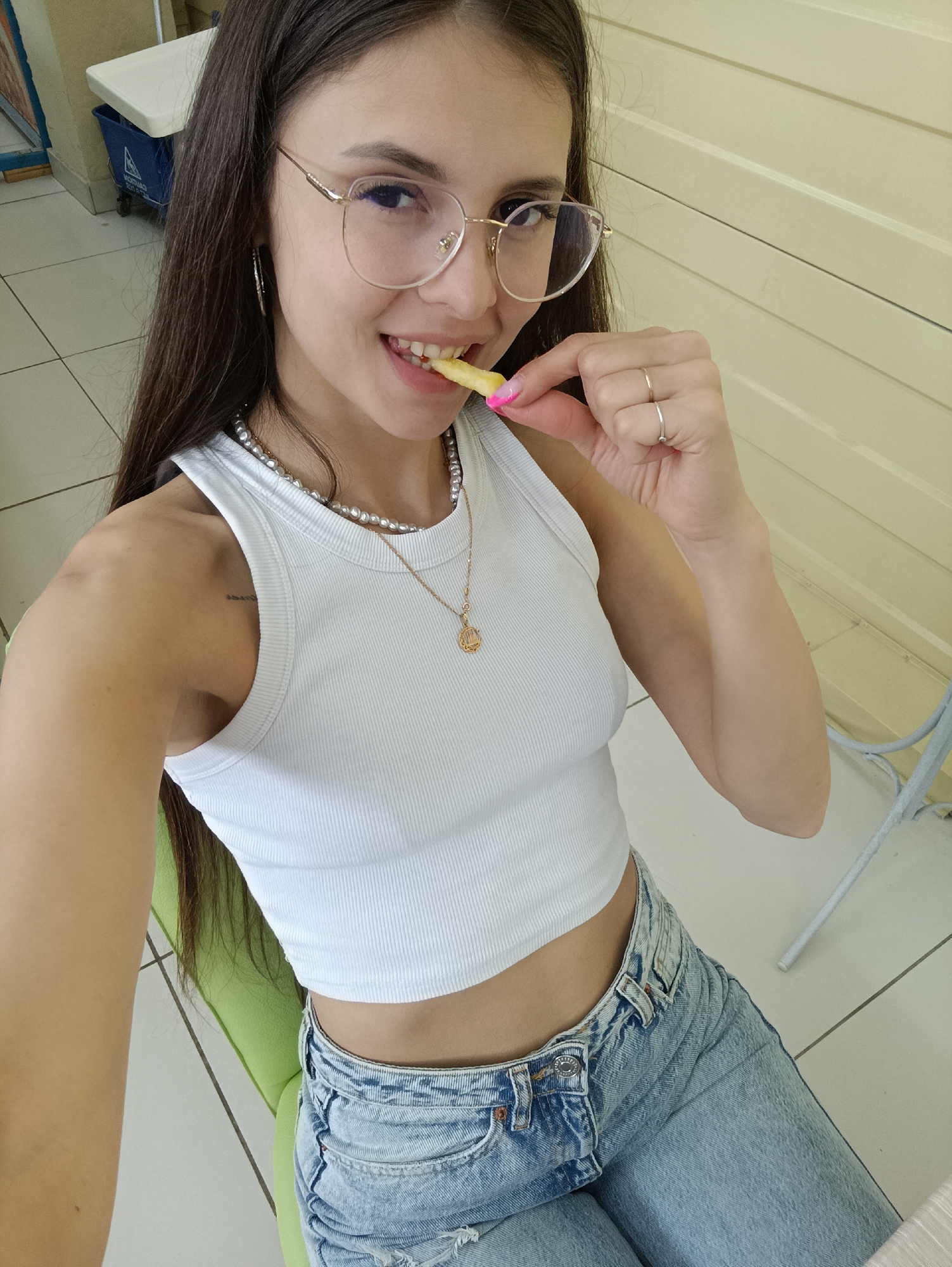 A young woman with long dark hair and glasses is shown from the chest up, sitting in a chair and eating a french fry. She is wearing a white ribbed crop top and blue denim jeans. She has on a pearl necklace and a gold pendant. Her nails are painted pink. The background is blurred but appears to be an indoor setting with light-colored walls and tiled floors. The lighting suggests it is daytime.