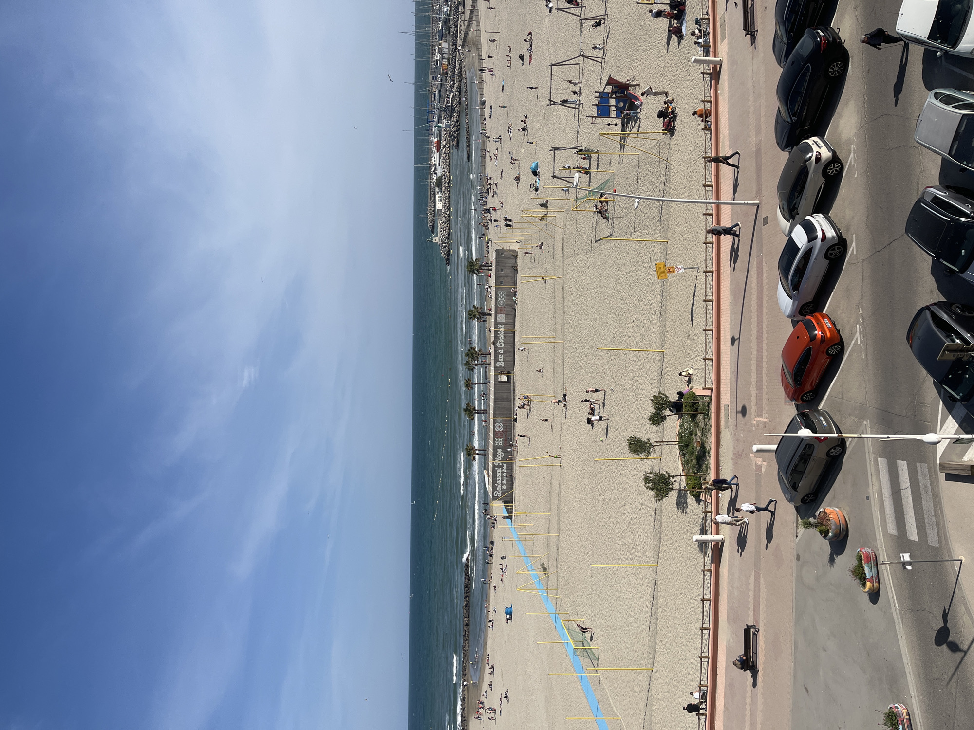 This aerial image captures a vibrant coastal scene, featuring a sandy beach, a busy harbor, a promenade, and a street with parked cars, all bathed in bright daylight under a clear blue sky with some wispy clouds. The setting is consistent with a Mediterranean beach town like Palavas-les-Flots, France.
The foreground is dominated by a wide, light-colored sandy beach populated by numerous individuals. People are seen relaxing, sunbathing, and walking along the shoreline. Children are engaged in play on a blue and yellow playground structure. A distinctive blue line traverses the sand, flanked by yellow poles, possibly delineating a specific area or path. Along the beachfront, two buildings house establishments, with visible text reading "Bar à Cocktail" and "Restaurant Plage Les pieds dans le sable." Several palm trees and other small trees are interspersed across the beach.
To the left, the calm, deep blue sea stretches out, with gentle waves breaking against the shore. A stone jetty or breakwater extends into the water, sheltering a harbor crowded with numerous docked sailboats and motorboats.
Separating the beach from the road is a paved promenade where a few people are walking. Adjacent to the promenade, a street is lined with parked cars of various colors, including black, white, orange, and grey. A pedestrian crosswalk is visible on the asphalt, and planters with greenery are placed along the roadside. Strong shadows indicate a time around mid-day or early afternoon, suggesting a warm and sunny environment. The overall scene conveys a lively, leisurely atmosphere typical of a popular beach destination.