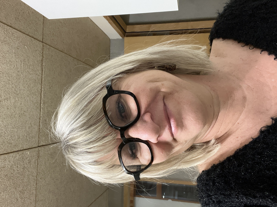 A blonde woman wearing black, square-framed glasses faces the camera. She has fair skin and blue eyes. The background features a beige wall with a visible linear pattern and a white surface near the top. Her hair is styled in a short, layered cut. She is wearing a dark, textured sweater. The image is rotated counter-clockwise.