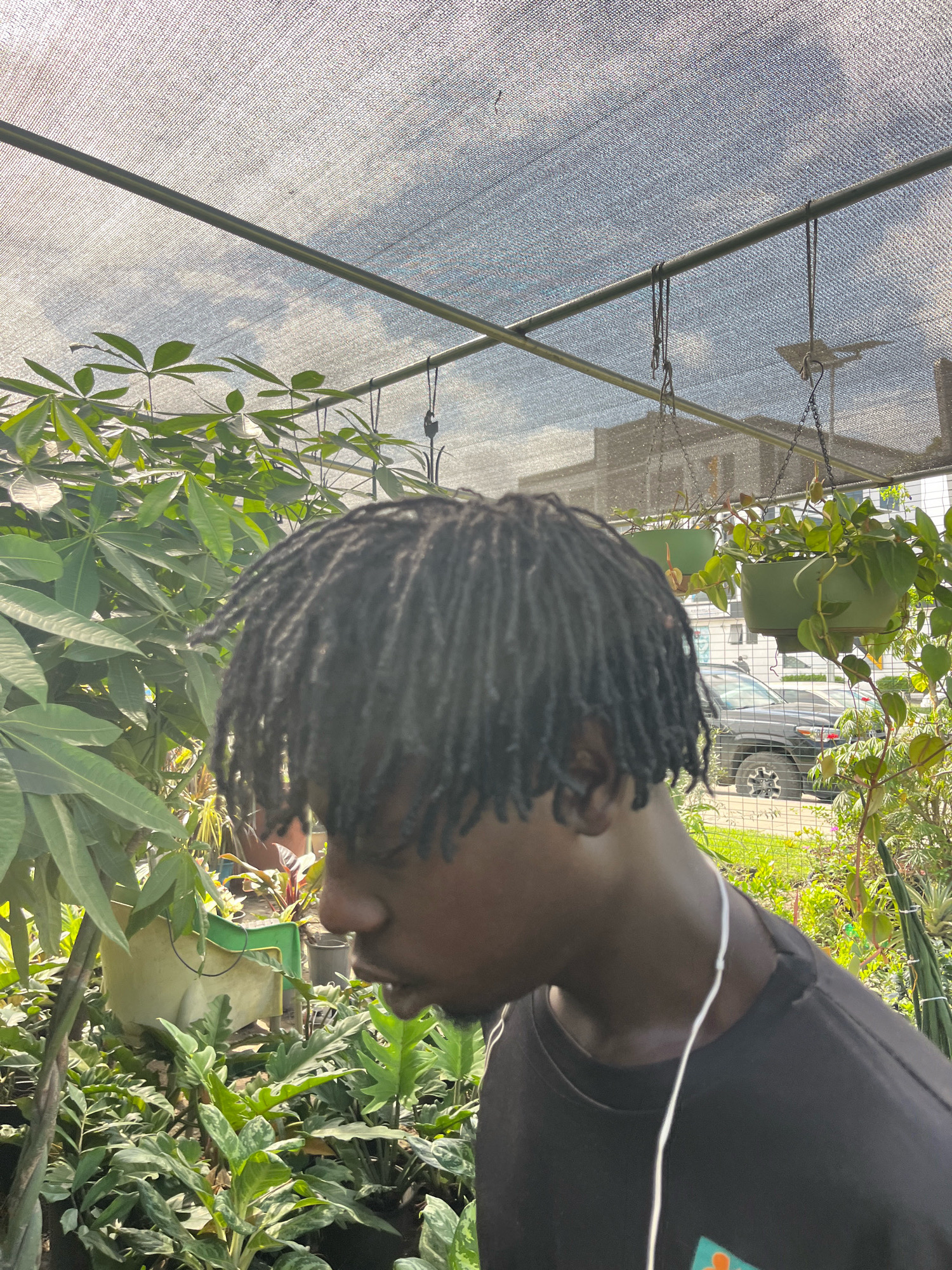 A young Black male with dreadlocks is shown in profile from the shoulders up, looking down and to his left. He is wearing a black t-shirt with a white cord visible. He appears to be in an outdoor plant nursery, with numerous plants of various sizes and types surrounding him. A translucent canopy is visible overhead, supported by metal poles, casting a dappled light. In the background, beyond the plants and canopy, a dark-colored SUV, a building, and some trees are discernible. The scene suggests a bright, possibly warm, day. The image is composed with the man's head and upper body as the primary focus, with the plants and the outdoor environment serving as the backdrop.