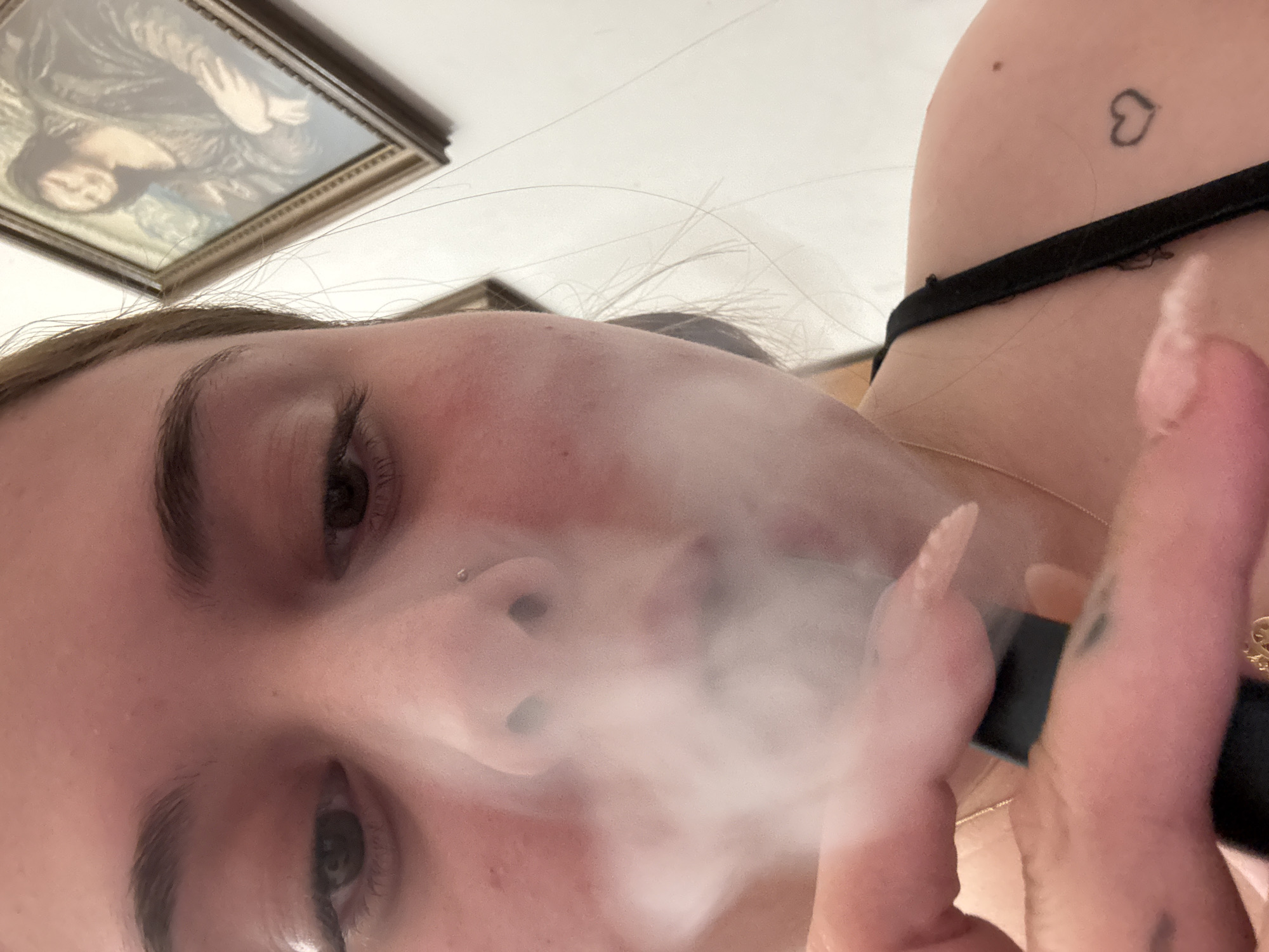 The image shows a person's face close-up, obscured by a cloud of vapor, possibly from a vape pen or e-cigarette which they are holding. The person has fair skin, dark eyes, and a nose piercing. They have long, light-colored nails. A black strap is visible around their neck. There is a heart tattoo visible on their chest. In the background is a framed painting on a white wall. There is no indication of the location being Troyan, Bulgaria.