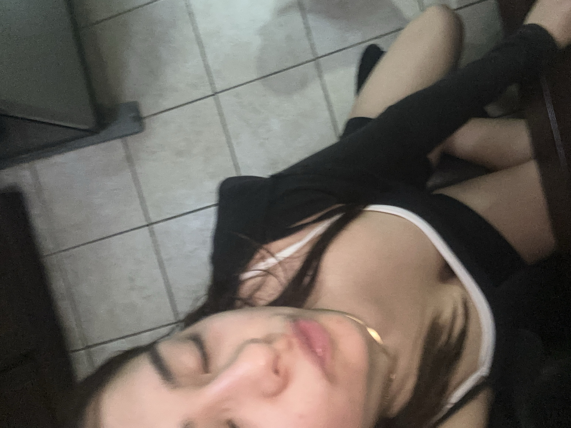 A young woman is lying on a tile floor in what appears to be a kitchen. She is wearing a white tank top and black leggings. Her face is close to the camera, showing closed eyes. The background features tile flooring and a section of a kitchen appliance. The location is presumably in Puebla, Mexico, but there is no definitive evidence to confirm this detail.