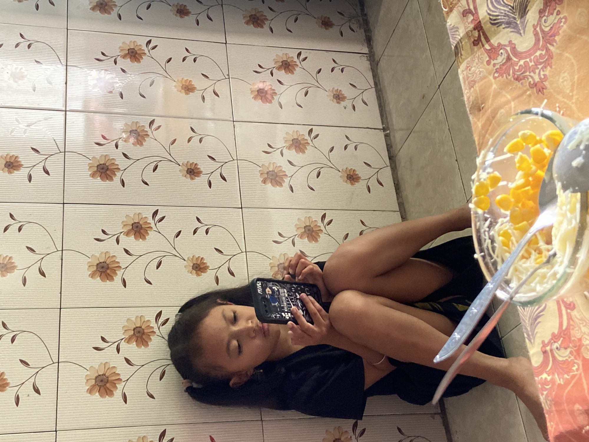 A young girl with dark hair in pigtails sits on the floor, facing left, and looks down at a smartphone in her hands. She is wearing a black t-shirt. Her legs are crossed, and her knees are bent. In front of her, on a table covered with a patterned cloth, is a glass bowl containing corn kernels and shredded white cheese. Two spoons are in the bowl. The background consists of a tiled wall with a floral pattern. The floor is tiled. The scene is likely indoors, possibly in a residential setting in Baron, Indonesia.