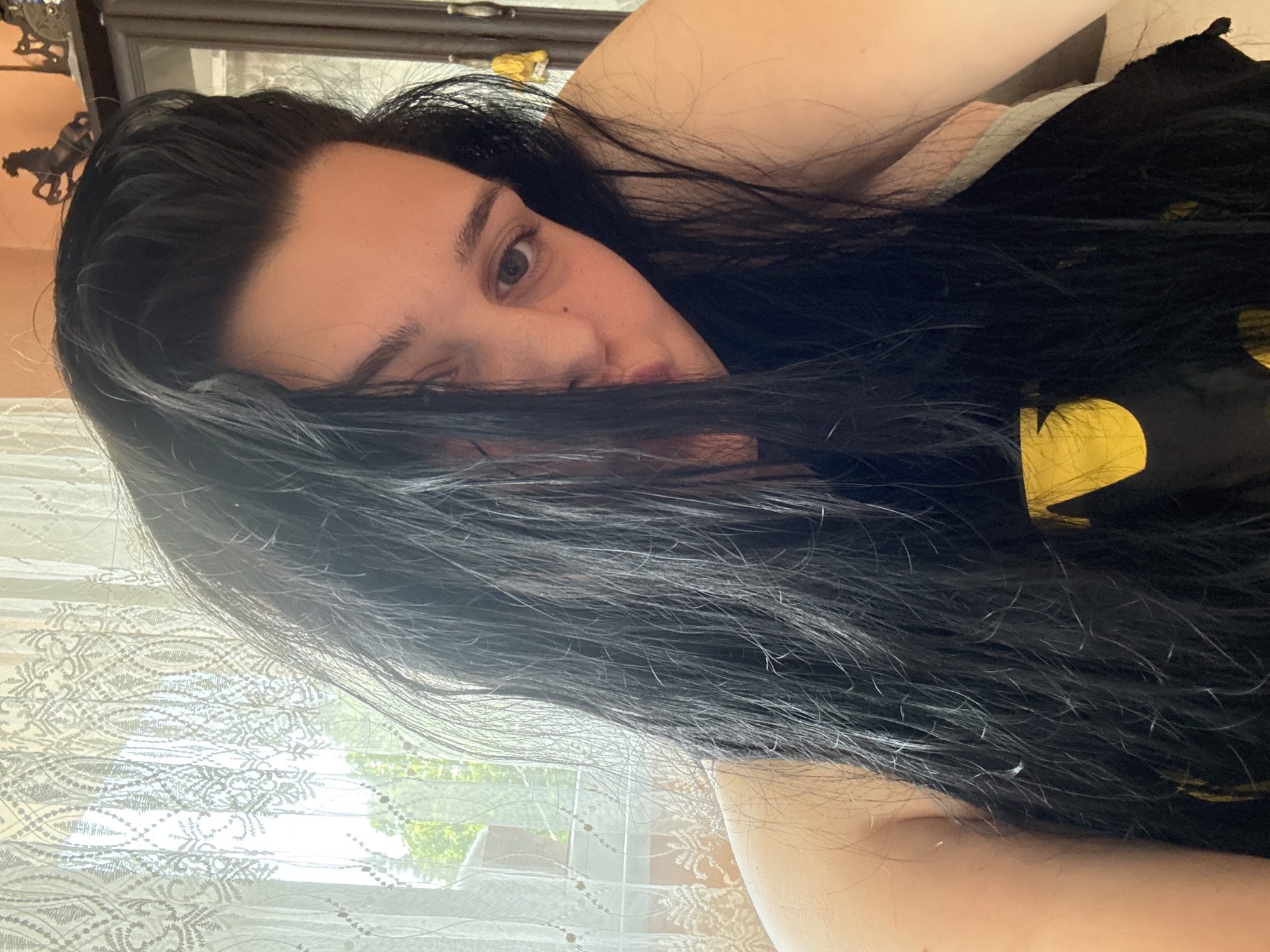 This media file presents a close-up, eye-level portrait of a person, likely a selfie, oriented at a 90-degree angle with the head on the left. The subject has long, dark, straight hair that partially covers their face, leaving one dark brown eye clearly visible. A small mole is visible on the cheek below the eye. The person has fair to medium skin tone and appears to be in a relaxed posture, possibly lying down.
They are wearing a black top, likely a t-shirt, with a distinct yellow design on the chest, possibly a stylized logo. The background features a window covered by a sheer, light-colored lace curtain with an intricate pattern. Bright daylight streams through the window, revealing green foliage and indistinct outdoor structures beyond, suggesting it is daytime. In the upper left corner of the image, a small decorative object, resembling a person riding a horse, is visible on a surface. The overall atmosphere is calm. No text is visible in the image. The location context is Rogosh, Bulgaria.