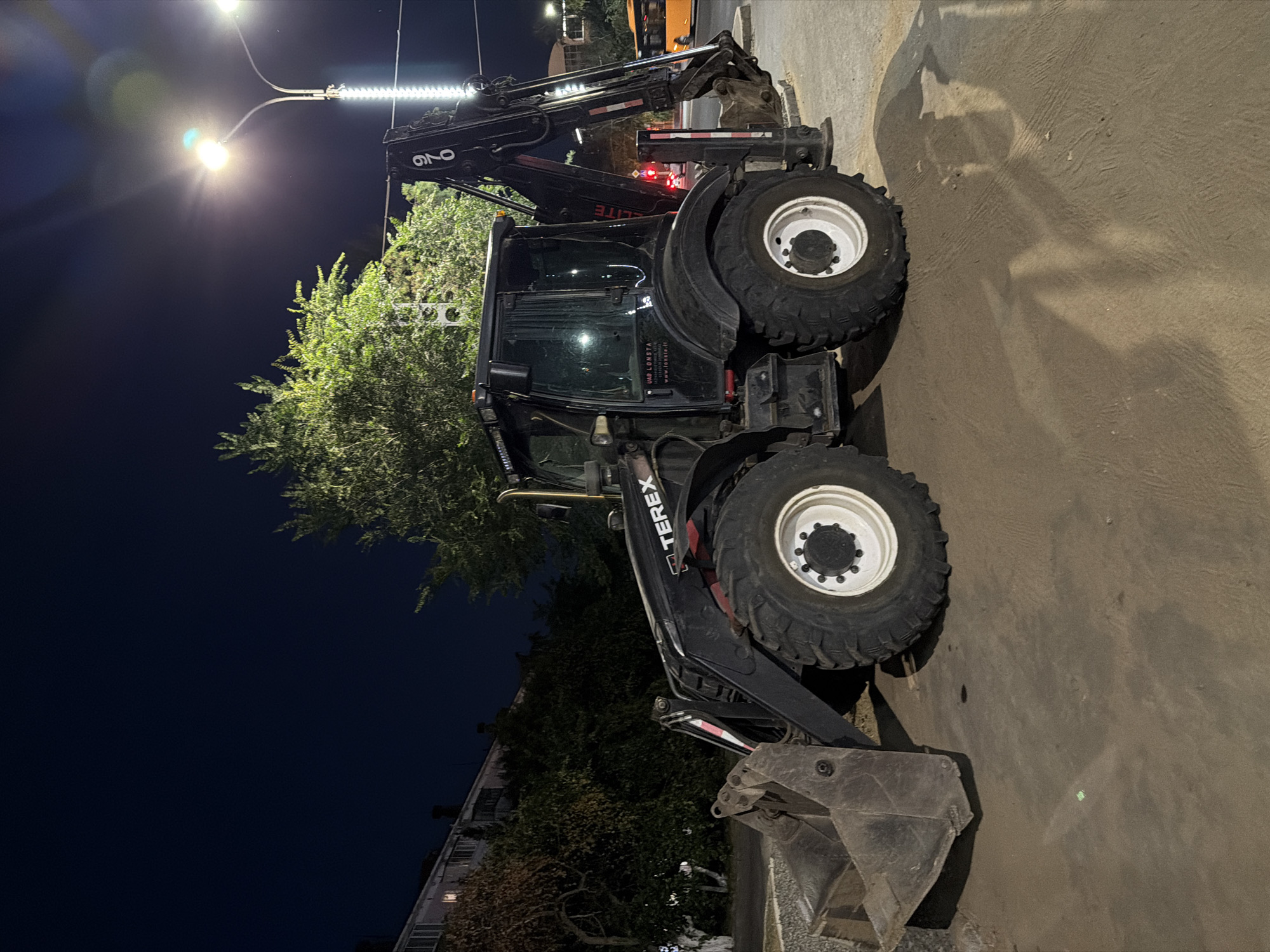A black Terex backhoe loader is parked on a dirt and asphalt surface at night. The backhoe is the primary subject, facing left and slightly towards the viewer, with its front loader bucket resting on the ground. The vehicle has large, knobby tires and a visible "TEREX" logo on its side. Behind the backhoe, a dense green tree is illuminated by streetlights. To the left, a multi-story building with visible windows is partially visible, suggesting an urban setting. In the background, a street with a traffic light and an orange bus are faintly discernible. The sky is dark, indicating nighttime. There are no people visible in the image. The scene appears to be in the city of Pavlodar, Kazakhstan, based on the presence of the specific markings on the backhoe, "UAB LONSTA www.lonsta.lt," which is a Lithuanian company that sells construction equipment. This suggests the backhoe might be in use or storage in a location where such equipment is operated.