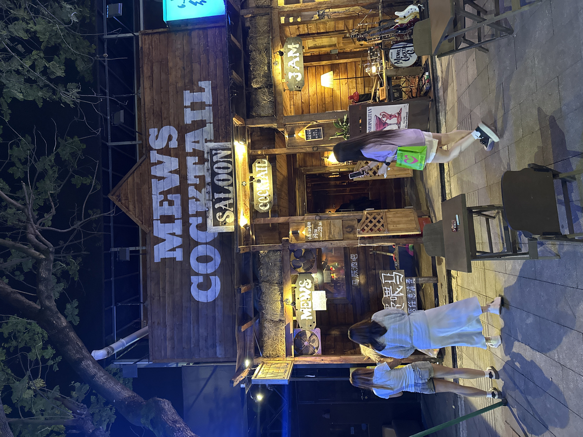 This nighttime image captures the exterior of a lively bar named "Mews Cocktail Saloon" in Chongqing High-tech Zone, China. The establishment features a rustic wooden facade with prominent white lettering spelling "MEWS COCKTAIL SALOON." Various smaller signs, some illuminated, also display "MEWS," "COCKTAIL," "JAM," and "3AM."
Four women are visible in the scene. In the foreground, two women stand on the paved sidewalk; one wears a light blue top and a long white skirt, while the other is in a white t-shirt and denim shorts. Further into the frame, a woman in a purple top and white shorts, carrying a bright green bag, walks past a table and chairs positioned outside the bar. Another person is partially visible inside the building.
The bar's entrance is adorned with a decorative wooden structure, hay bales, and several fans. Inside, musical instruments including guitars and a drum set are displayed, suggesting live performances. Tables and chairs are arranged on the sidewalk for outdoor seating. The setting is illuminated by the bar's lights and overhead streetlights, casting shadows on the pavement. A large tree with dark foliage is on the left, contrasting with the warm glow from the bar.
Visible Chinese text includes "小馬上班時間 19:30 - 23:30" (Xiao Ma's working hours 19:30 - 23:30), "马厩酒吧" (Mews Bar), "喂马" (Feed Horse), and "本店任意消费" (Any consumption in this store). Other texts like "BOULANEUR," "VERITASEVE," and "SMOPOLITAN" (likely part of "COSMOPOLITAN") are also present on posters or signs. The atmosphere appears casual and inviting for an evening out.