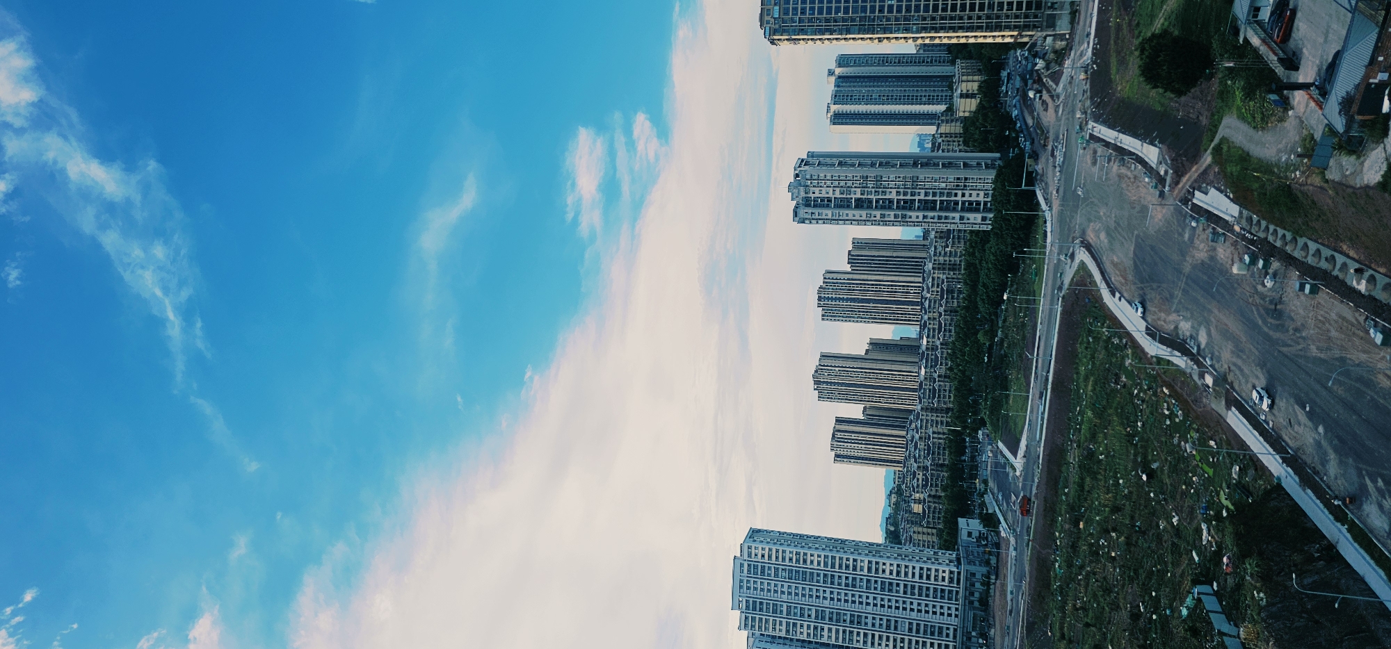 The image depicts a developing urban landscape in Jiulongpo District, China. In the background, a cluster of tall, modern apartment buildings dominates the skyline, with some smaller residential structures visible behind them. The buildings are characterized by their repetitive window patterns and varying heights. In the midground, a dense green belt of trees separates the residential areas from a construction site. The foreground shows an unfinished road and adjacent undeveloped land. Several white cars are present on the road, and a single orange truck is visible in the distance. The sky above is a vibrant blue with wispy white clouds. The overall scene suggests a city undergoing expansion and construction.