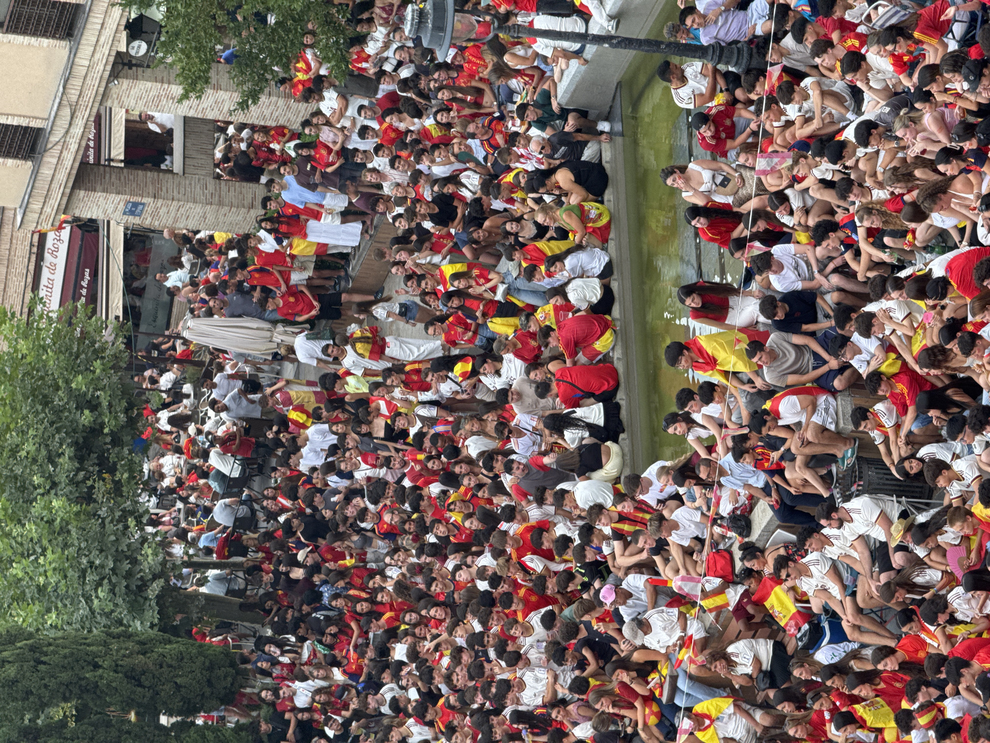 A crowd of people, many wearing red and yellow, the colors of the Spanish flag, are gathered in an outdoor urban setting. They are standing and sitting on tiered stone steps surrounding a shallow water feature or pool. The people are densely packed, with some individuals draped in Spanish flags. Buildings, some with awnings and signage, are visible in the background, suggesting a public space or plaza. Trees and foliage are present, indicating an outdoor environment. The scene appears to be a gathering for a public event, possibly a sporting event or celebration, in Las Rozas de Madrid, Spain.
