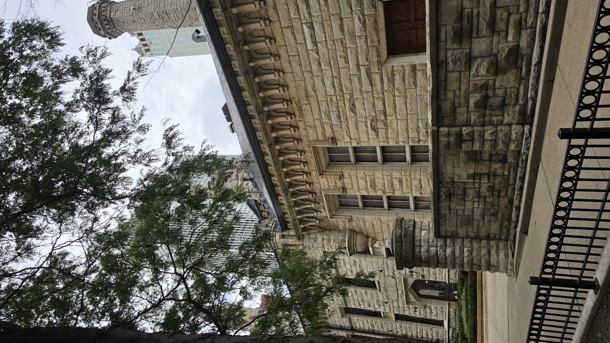 A low-angle exterior medium shot captures a stone building in Chicago, Illinois. The building's facade is constructed of light-colored, rough-hewn stone blocks. The upper portion of the building features decorative stone corbels and a dark, sloped roof. Two tall, narrow windows with multiple panes are visible on the facade, as is a wooden door with a paneled design. In the foreground, a low, rough-hewn stone wall runs horizontally across the image. A black metal fence with circular accents borders the sidewalk in front of the wall. A large tree with dense green foliage partially obscures the upper left portion of the frame, revealing a glimpse of a modern, glass-windowed skyscraper behind it. The sky is overcast and gray.