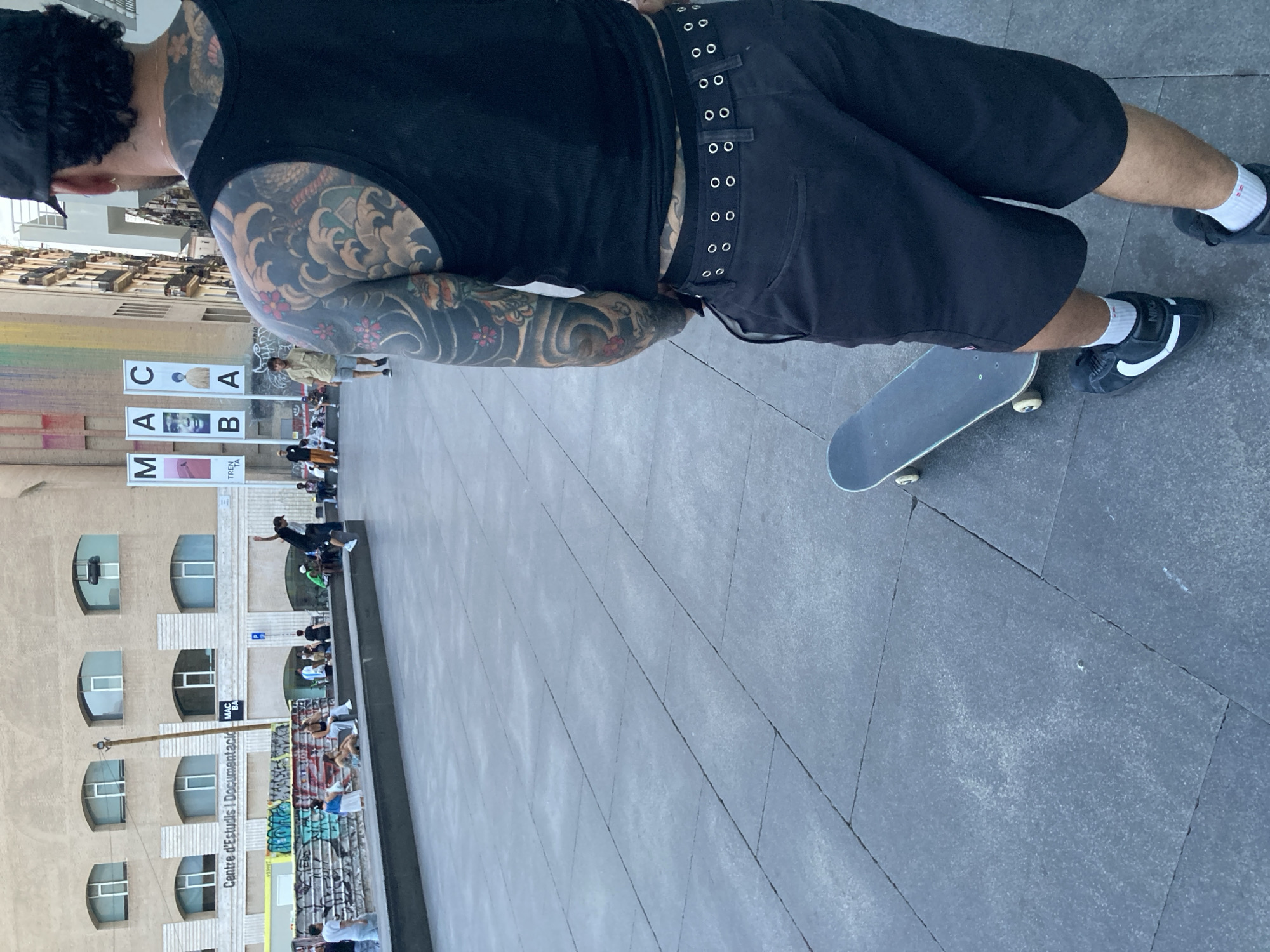 In Barcelona, Spain, a man with extensive tattoos on his arms and back stands facing away from the camera, wearing a black tank top, black shorts, and black sneakers. He is holding a black skateboard, which rests on the grey tiled ground in front of him. In the background, several people are visible, some sitting on a concrete ledge, others standing and walking. Behind them, a building displays banners with the letters "MACBA," indicating the Museum of Contemporary Art of Barcelona. The area appears to be a public plaza.