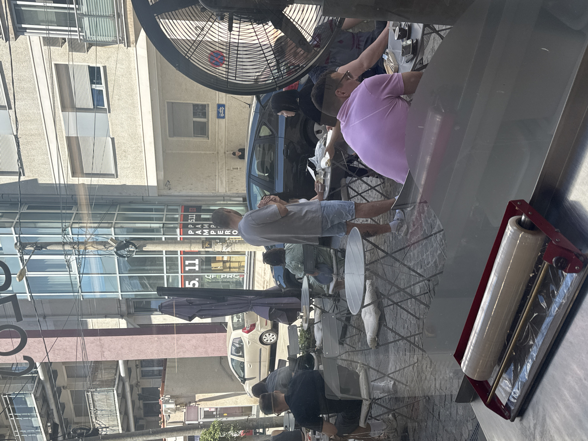 The image shows an outdoor cafe scene, likely on a busy street in Vracar Urban Municipality, Serbia, as suggested by architectural styles and store signage. Several people are seated at small tables, eating and drinking. A man wearing a pink shirt is seated at one table, while another man in a gray shirt stands nearby. Other patrons are visible in the background. A large fan is suspended above the seating area. Through the reflection of the window, buildings and street signs are visible, including one that reads "5. 11,". A white car is parked on the side of the street. In the foreground, inside what appears to be a deli or eatery, is a plastic wrap dispenser.