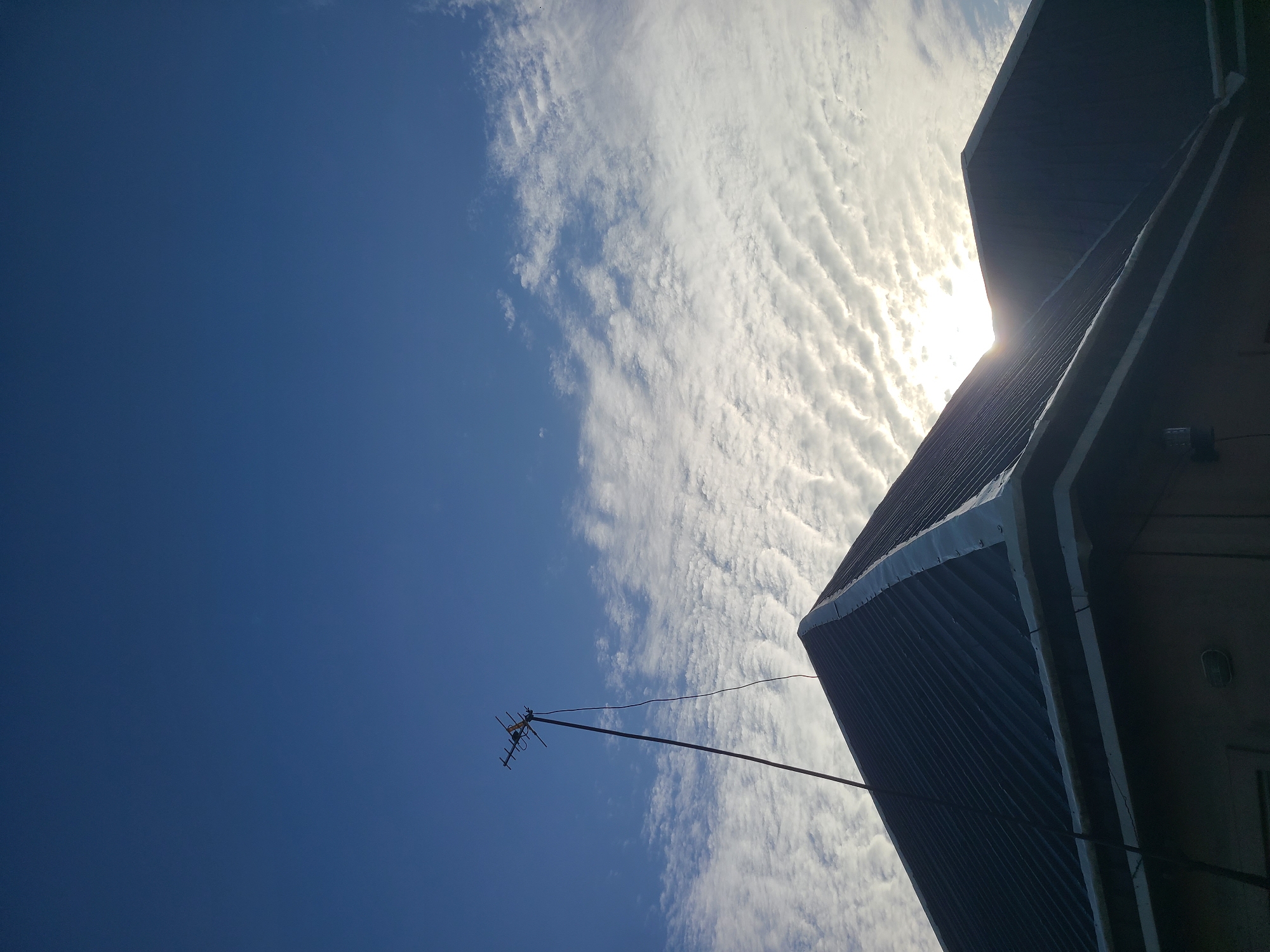 The image shows a dark, corrugated metal roof of a building against a bright blue sky with scattered white clouds. A tall, slender antenna with multiple elements is mounted on a pole that extends from the peak of the roof, reaching into the sky. A dark cable runs from the antenna down to the roof. The sun is partially visible behind the clouds, casting a bright glow and creating lens flare. The scene appears to be an exterior view of a residential or commercial building in Kuru Karama, Nigeria.