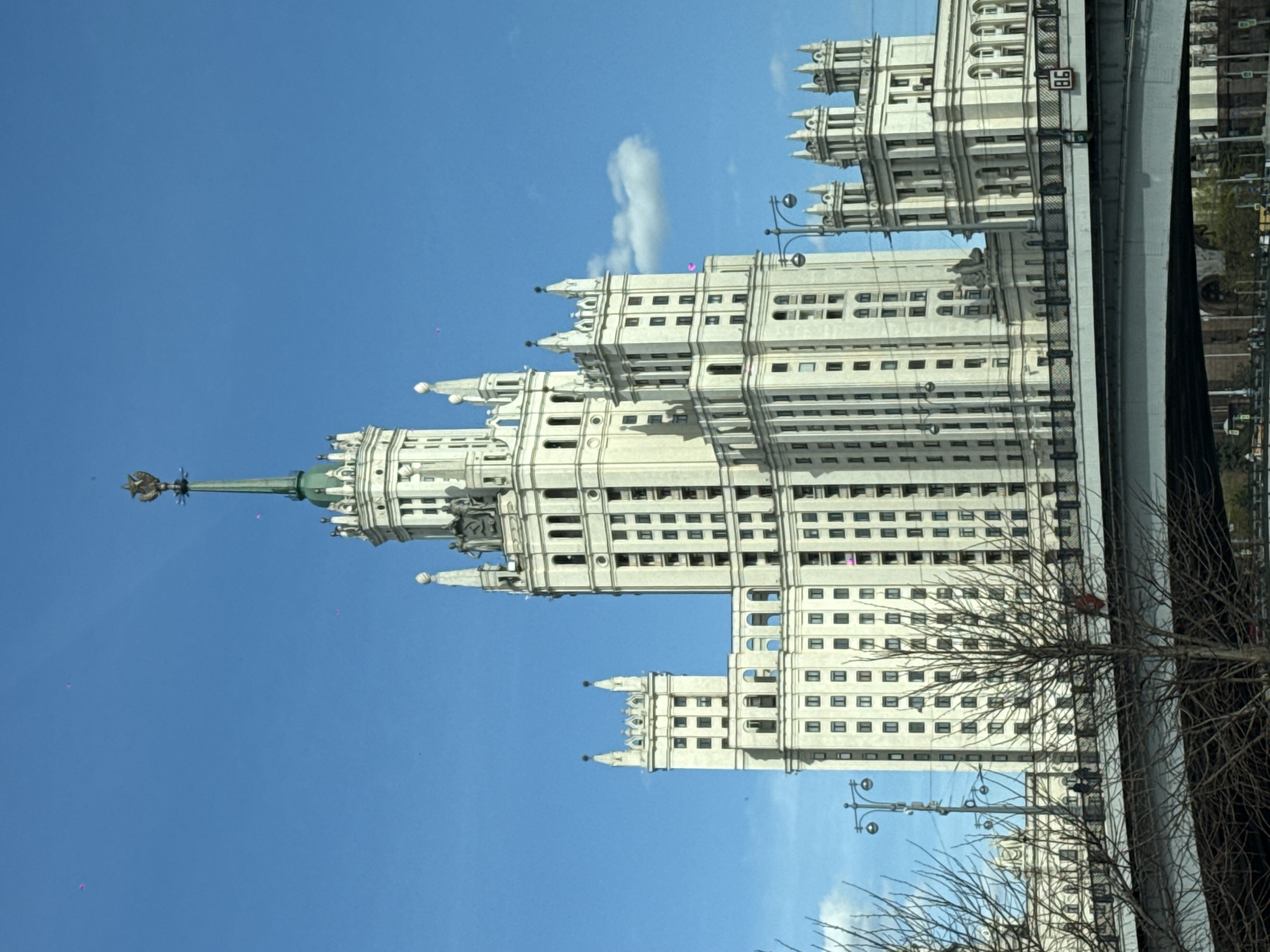 The image shows the Kotelnicheskaya Embankment Building, one of the "Seven Sisters" Stalinist skyscrapers in Moscow, Russia. The building is a large, white, ornate structure with multiple tiers and a spire. The sky is blue with scattered clouds. Part of a bridge and some bare tree branches are visible in the foreground.