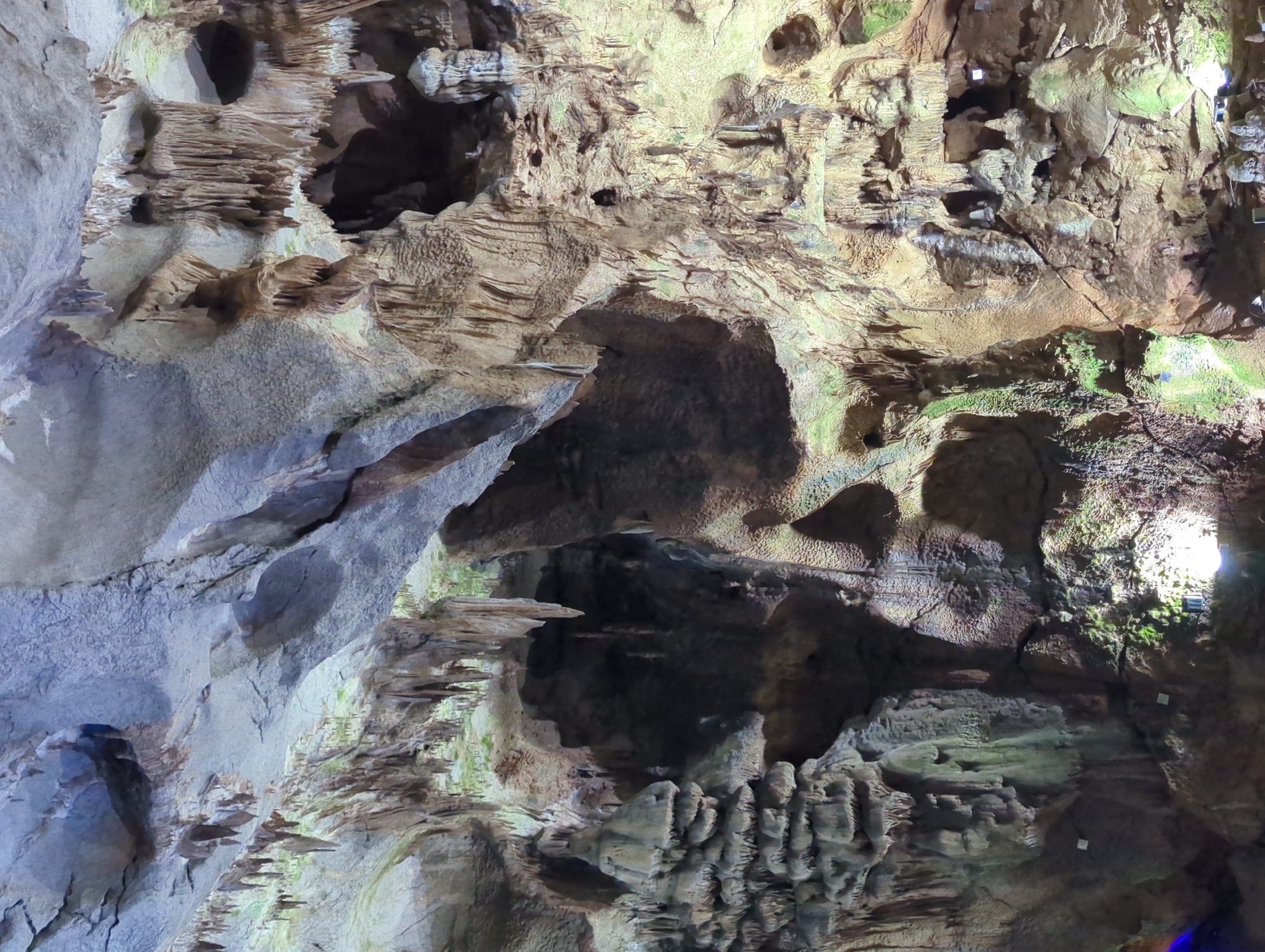 The image shows the interior of Ledenika Cave, located near the city of Vratsa, Bulgaria. The cave walls and ceiling are adorned with numerous stalactites and stalagmites, creating a complex and varied geological formation. Some of these formations are illuminated by artificial lights, casting shadows and highlighting the textures of the rock. There are no people visible in the image. The environment is that of a natural underground cave, characterized by its rock formations, darkness, and the presence of mineral deposits.
