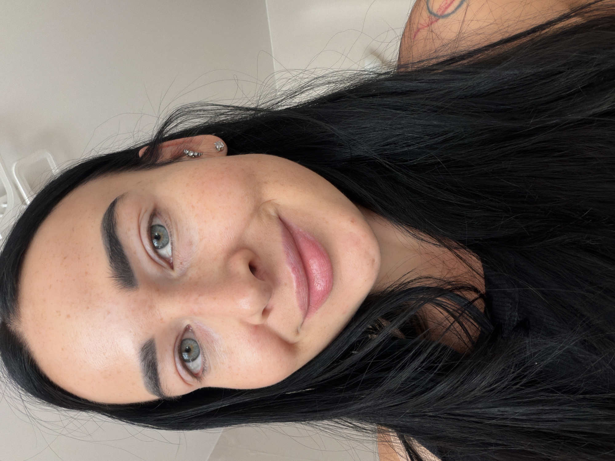 A selfie shot of a woman with fair skin, freckles, dark hair, and light-colored eyes, taken indoors. She has piercings in her left ear and a tattoo on her upper right shoulder. The background appears to be a white or light-colored wall. The image is oriented with the subject's head tilted to the left. There is no indication of the location being in Saint Petersburg, Russia, or any specific city.