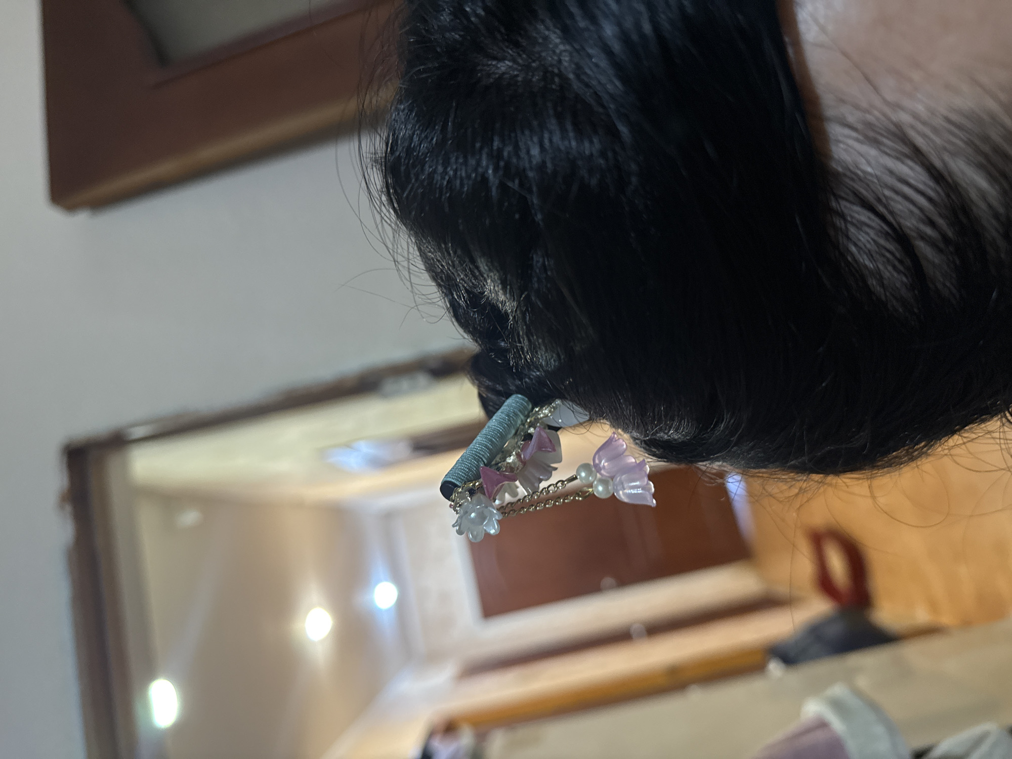 The image shows a close-up of a person's dark hair styled with a decorative hairpiece. The hairpiece is a blue chopstick with dangling charms that include small pink and purple bell-shaped flowers and white flower-shaped beads, along with pearls. The background is blurred, showing what appears to be an indoor hallway with doors and lighting fixtures. There are no discernible elements in the image that would allow for the identification of a specific location in the city of Dnipro, Ukraine.