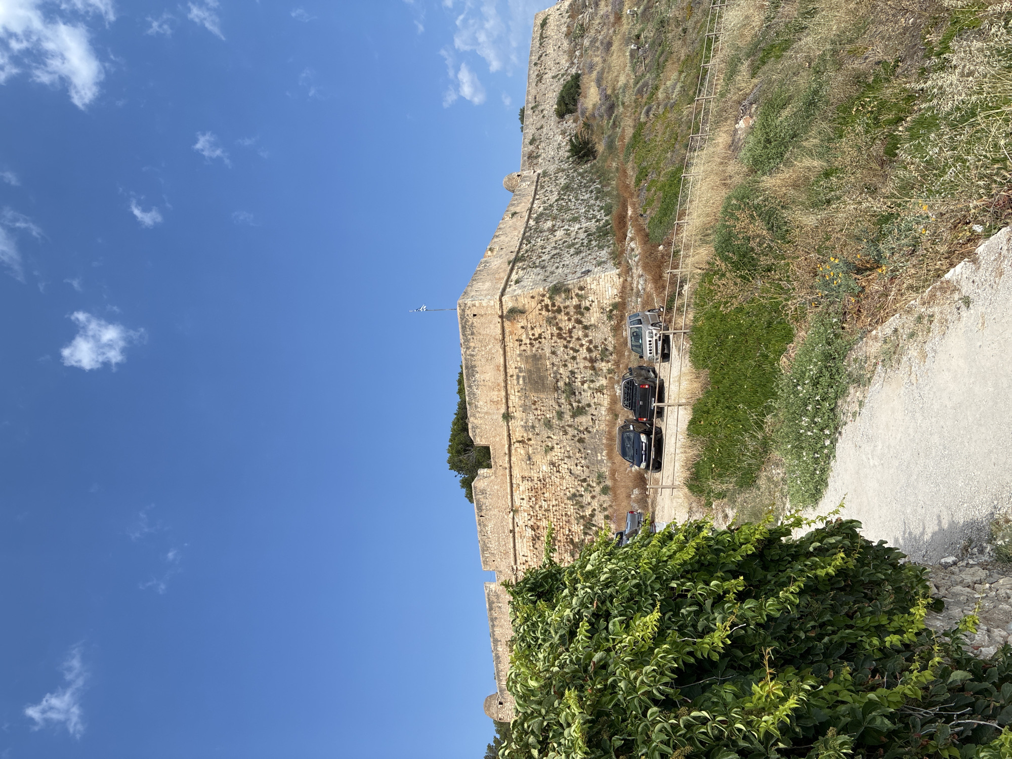 The image shows a view of a large stone fortification under a clear blue sky with scattered white clouds. The fortification appears ancient, constructed of weathered stone blocks. A pathway runs along the base of the fortification, with several vehicles parked on it. These include a silver SUV, a black SUV, and a dark-colored sedan. Lush green vegetation, including dense vines, grows at the bottom of the image, contrasting with the dry, grassy slopes of the fortification's exterior. A metal railing runs along the edge of the pathway, separating it from the natural terrain. A flag, possibly the Greek flag, is visible on a flagpole near the top of the fortification. The scene suggests a historical site, likely a castle or fortress, on a sunny day, possibly in the late morning or early afternoon given the bright sunlight and clear skies. The location appears to be a tourist attraction, given the presence of parked vehicles.