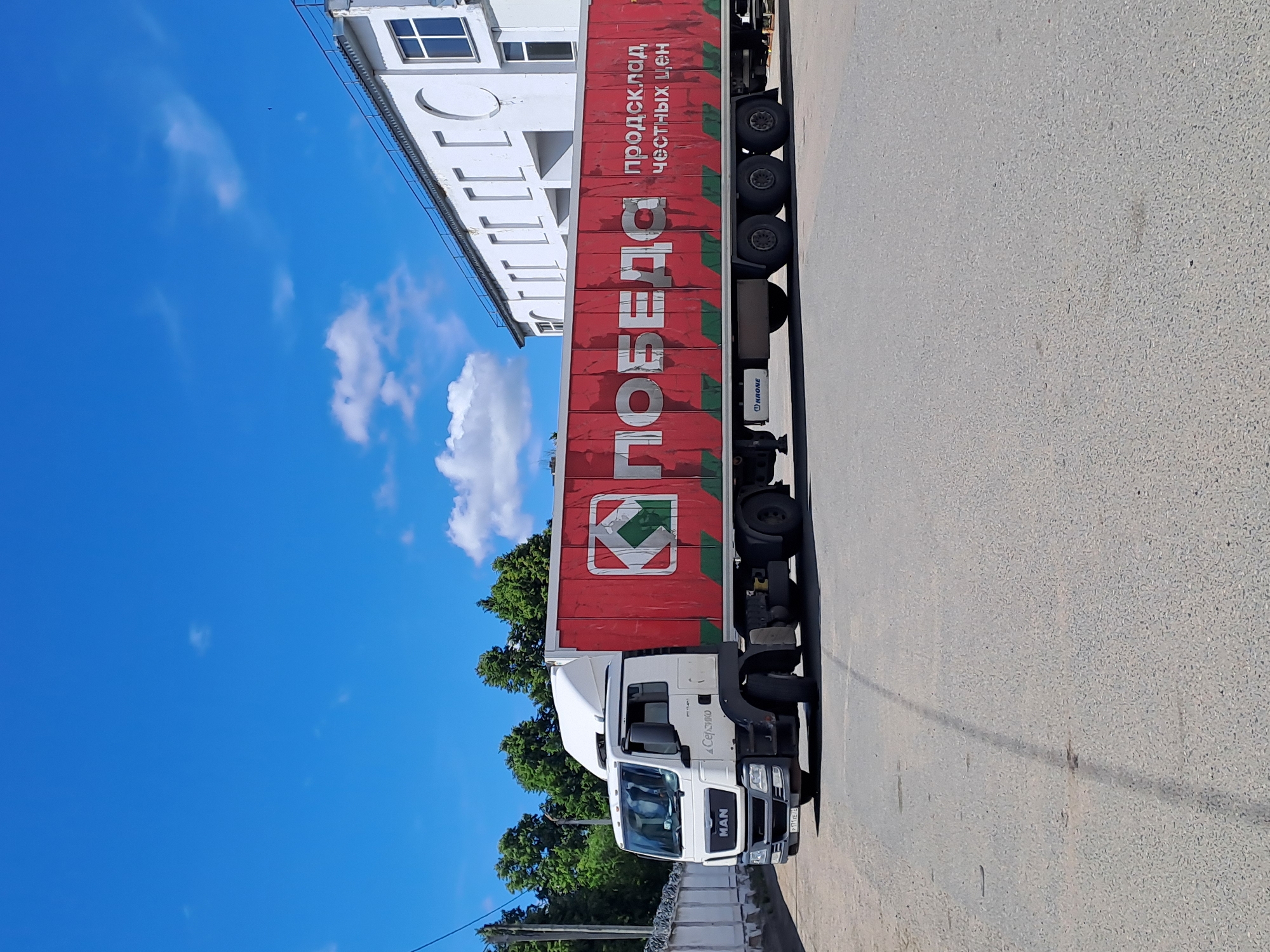 A white MAN truck with a large red trailer is prominently featured, parked on an asphalt road in Yalga, Russia. The red trailer is extensively branded with white Cyrillic text. Large letters spell "ПОБЕДА" (Pobeda), which translates to "Victory." Above this, smaller text reads "ПРОДСКЛАД ЧЕСТНЫХ ЦЕН" (Prodsklad Chestnykh Tsen), meaning "Grocery Warehouse of Honest Prices." A red and green geometric logo is also visible on the trailer, and the brand "Krone" is written on the trailer's chassis.
The truck's cab has "MAN" visible on its grille and "Сержио" (Sergio) written vertically on its side. A partial license plate "111 XE 66" can be seen. To the left of the truck is a multi-story white building with several rectangular windows and decorative architectural elements. Behind the truck and building, lush green trees are visible against a vibrant blue sky dotted with scattered white clouds, indicating a bright, sunny daytime. The asphalt road in the foreground appears a light grey. The scene depicts a commercial vehicle in an urban or industrial setting.