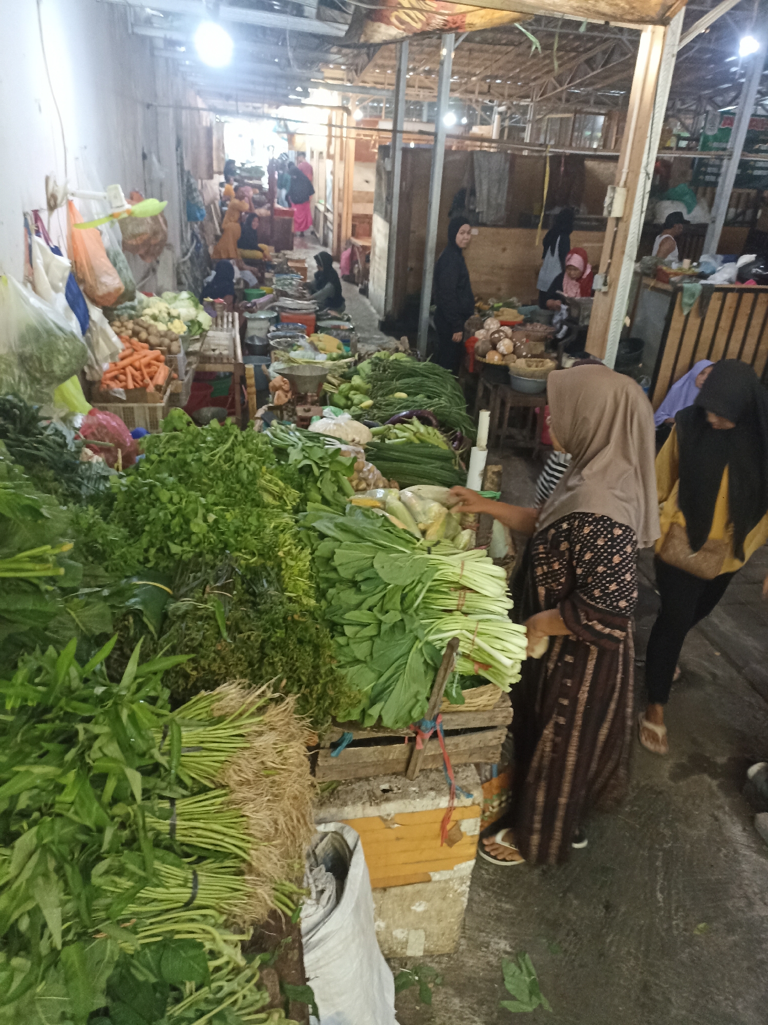 The image depicts an indoor market scene in Kelurahan Kepatihan, Indonesia, likely a wet market. The foreground is dominated by a large display of fresh vegetables, including bundles of green leafy produce with visible roots, carrots, and other greens. Several people are present, mostly women, some of whom appear to be vendors or shoppers. One woman in the center, wearing a brown hijab and a patterned dress, is interacting with the vegetables. Another woman in a black hijab and yellow top stands to her right. In the background, more market stalls and people are visible, along with wooden structures and what appears to be corrugated metal roofing, suggesting a rustic or traditional market setting. The lighting is somewhat dim and appears to be from overhead artificial sources.