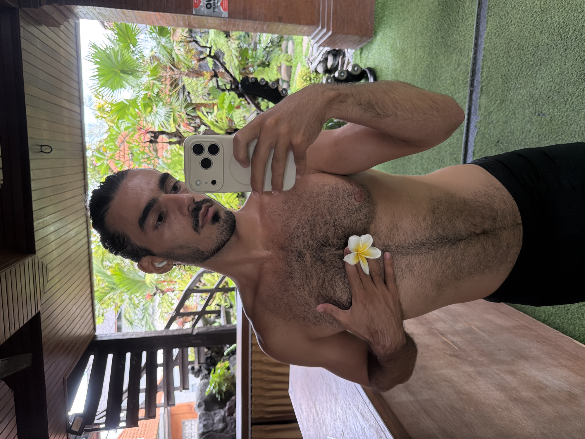 A shirtless man takes a mirror selfie in a tropical setting. He is positioned slightly left of center, looking into a light-colored smartphone held by his right hand, reflecting the scene. His dark hair is styled back, and he has a beard and mustache. An earbud is visible in his left ear. A white and yellow plumeria flower rests on his chest, with his left hand gently touching it. He has visible chest and arm hair.
The reflected background behind him reveals a lush outdoor environment with vibrant green palm trees and other tropical foliage under a bright sky, suggesting daytime. Indoor elements reflected include a green, textured floor (likely a gym mat or artificial turf), some gym equipment (appearing as dumbbells or weights) towards the middle right, and warm-toned wooden structures like paneling, railings, and what looks like a staircase. A wooden table or bench is visible in the lower right foreground.
The combination of tropical plants, wooden architecture, and the plumeria flower strongly indicates a location in a tropical region, consistent with North Kuta, Indonesia. The setting appears to be a fitness area or gym that is integrated with or overlooks natural surroundings.
In the upper left corner of the reflected background, a red object with reversed and partially visible white text can be seen, though the specific words are illegible due to the reflection and blur.