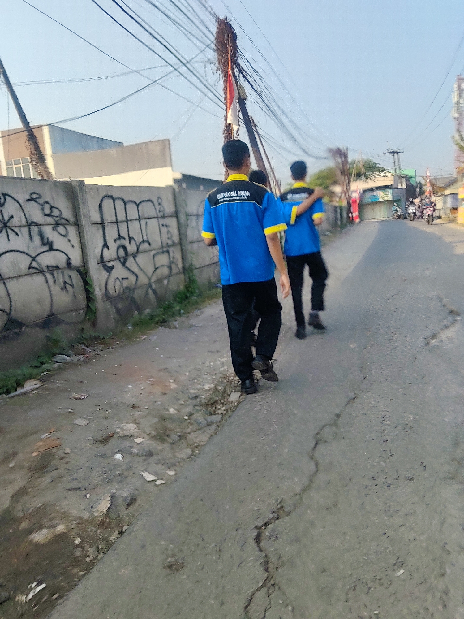 Two young males, likely students wearing matching blue polo shirts with yellow accents and black pants, walk away from the camera down a paved street. The shirt of the male in the foreground has text that appears to read "SMK GLOBAL MURIA" and a web address. The male in the background has his arm around the shoulder of the male in the foreground. They are walking on the right side of the road. The street appears to be in a semi-urban area of Mekarmukti, Indonesia, as indicated by the presence of buildings, overhead utility wires, and what appear to be motorcycles and scooters in the distance. To the left of the road is a concrete wall covered in graffiti. The ground on the left side of the road is unpaved and appears to be dirt with some scattered debris. The sky is clear and blue, suggesting it is daytime.