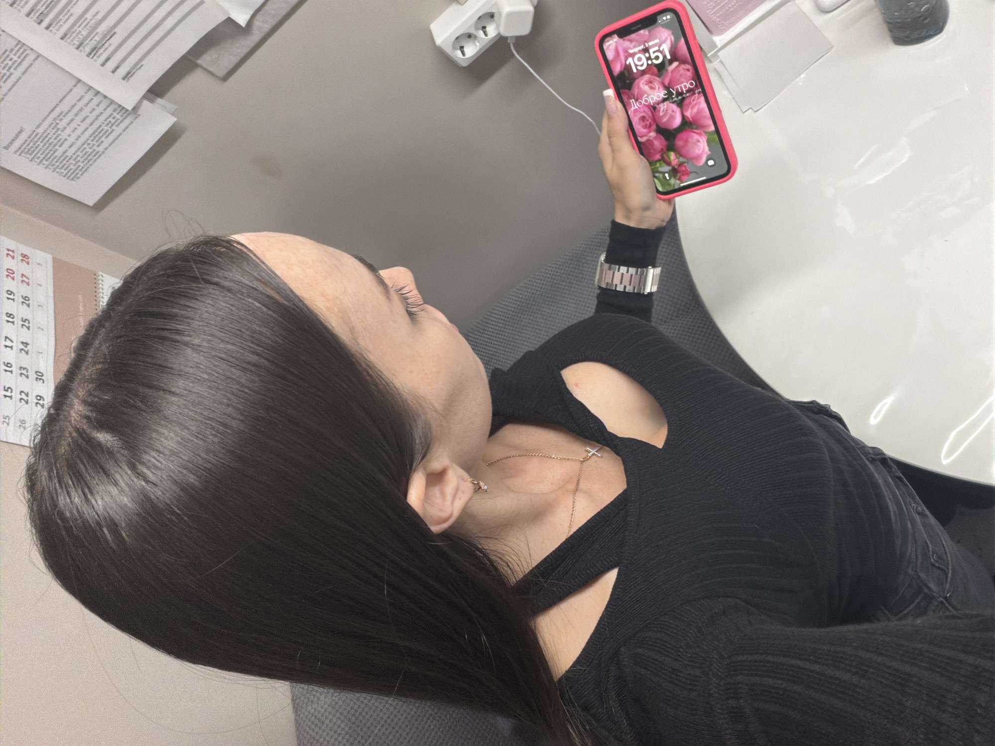 This media file depicts a close-up, high-angle shot of a woman from the shoulders up, taken indoors. The woman has long, dark hair and is wearing a black ribbed long-sleeved shirt with a cutout at the neckline. She is holding a pink-cased smartphone in her right hand, which is displaying a lock screen with the time "19:51" and the Russian text "Доброе утро" (Good morning) below an image of pink roses. The phone is plugged into a white electrical outlet.
The setting appears to be a desk or office environment. To the left of the woman, papers are stacked on a surface. A calendar with dates visible is also present to her left. To the right, a white table surface reflects light. A glimpse of a glass containing a dark liquid is visible on the far right. The overall lighting suggests an indoor setting, possibly late afternoon or evening, given the time displayed on the phone, despite the "Good morning" text on the screen. The woman's expression is not clearly visible due to the angle and focus. There's a delicate cross necklace visible on her neck.