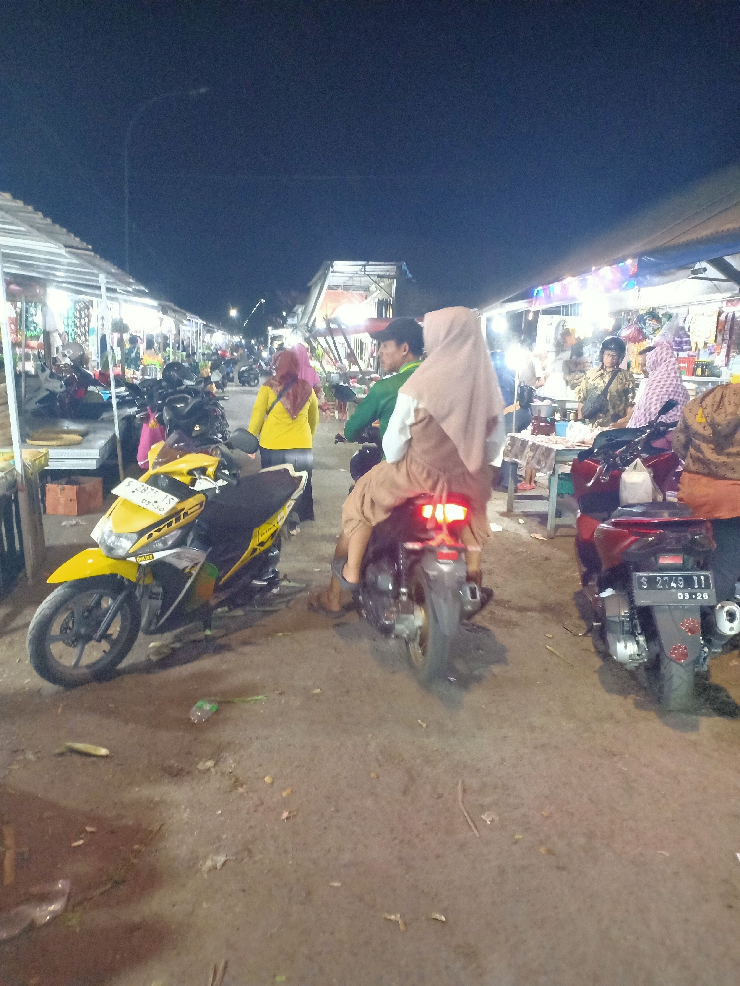 A nighttime outdoor market scene in Semanding, Indonesia, features a central street lined with vendor stalls on both sides, illuminated by artificial lights. The ground is a dusty, dirt-and-pavement mixture, with some scattered debris. Two motorcycles are prominent in the foreground: a yellow scooter on the left and a darker motorcycle on the right. Several people are present, some on foot and others on the motorcycles. On the yellow scooter, a person is seated, facing away from the camera. The darker motorcycle has two individuals on it, also facing away from the camera; the rider is wearing a helmet, and the passenger is covered in a light-colored headscarf and dress. Other individuals are visible in the background, browsing at the stalls. The stalls themselves display various goods, suggesting a commercial environment.