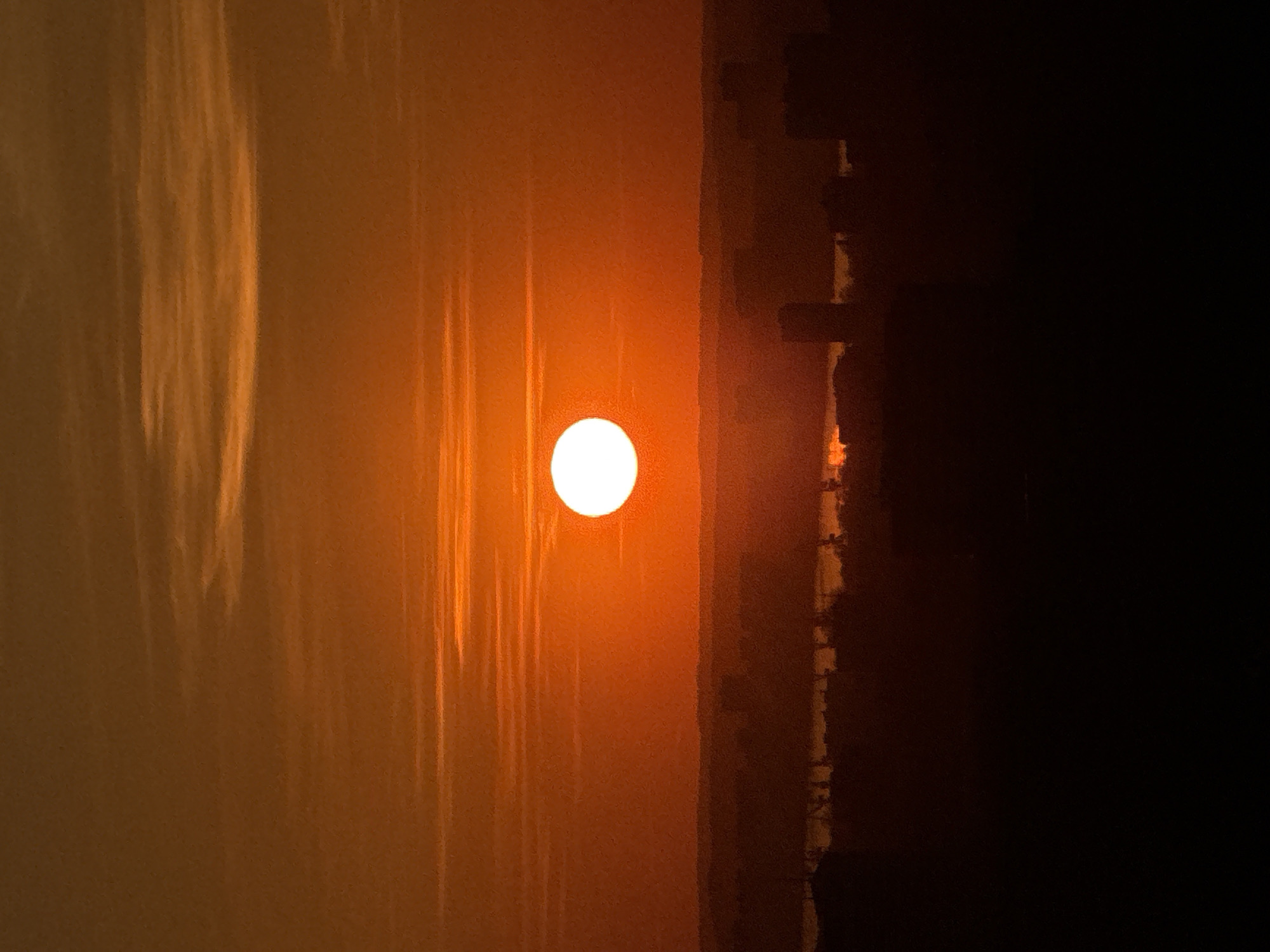 An eye-level shot shows an orange sun in a hazy sky, with a dark structure, possibly a building, obscuring the right portion of the frame, with the location appearing to be Perm, Russia.