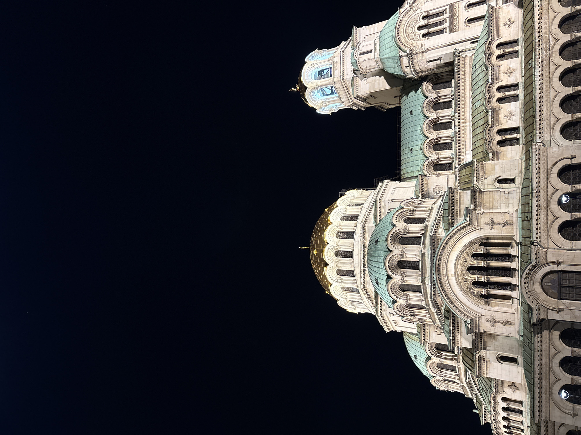 The image captures the Alexander Nevsky Cathedral in Sofia, Bulgaria, at night, brightly illuminated against a dark, clear sky. The grand Neo-Byzantine structure occupies the right side of the vertical composition, showcasing its majestic architecture. Multiple domes are visible, with several featuring a distinctive verdigris patina from oxidized copper, while the largest, central dome gleams in gold, reflecting the artificial light.
The cathedral's facade is crafted from light-colored stone or stucco and is richly adorned with intricate details. These include numerous arched windows, decorative columns, sculptural elements, and geometric patterns. The artificial lighting dramatically highlights the textures and three-dimensional qualities of the building, emphasizing its complex construction and ornate embellishments. No individuals or specific events are depicted, placing sole focus on the architectural beauty of the landmark. The complete absence of clouds against the deep black background confirms a clear night. There is no visible text.