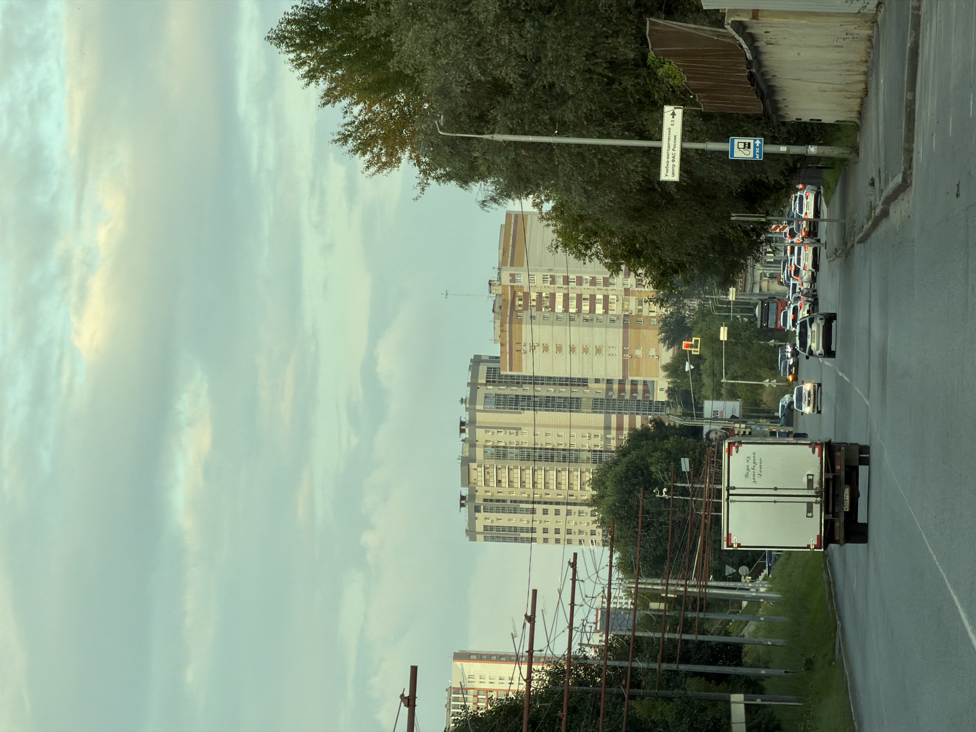 The image shows a street scene in an urban environment, likely during daylight hours with overcast skies. A paved road runs horizontally across the lower portion of the frame, with multiple lanes of traffic. Several vehicles are visible on the road, including a white box truck in the foreground, followed by a line of cars. On the right side of the road, there is a sidewalk and a wall constructed of rough, light-colored stone or concrete, topped with a wooden fence. A tall street light stands beside the sidewalk. On the left side of the road, a row of utility poles with overhead wires suggests a public transportation system, possibly for trams or trolleybuses. In the background, large apartment buildings, characteristic of urban residential areas, rise above the trees. The sky is filled with soft, diffused light, indicating cloudy conditions. A sign on the street light pole is visible, bearing text in Cyrillic.