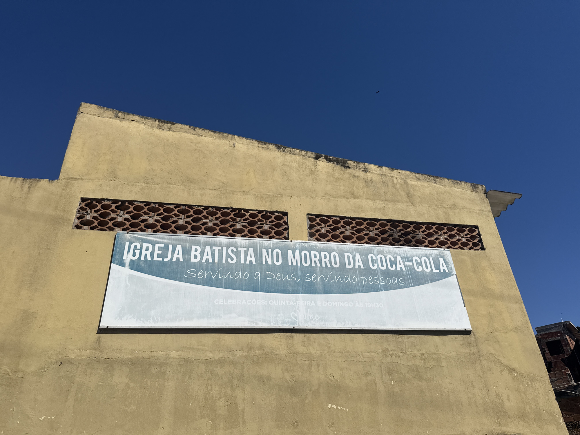 A white sign with blue lettering is mounted on a weathered, yellowish-tan building under a clear blue sky. The sign reads "IGREJA BATISTA NO MORRO DA COCA-COLA" followed by "Servindo a Deus, servindo pessoas." Below this, it states "CELEBRAÇÕES: QUINTA-FEIRA E DOMINGO ÀS 19H30." Two rectangular openings with decorative red brick patterns are visible above the sign. The building appears to be in a somewhat deteriorated state, with visible cracks and discoloration on its facade. In the upper right portion of the image, a glimpse of a brick structure, possibly another building under construction or in disrepair, is visible. The overall scene suggests a location in a town or city, likely Arraial Do Cabo, Brazil, given the context provided.