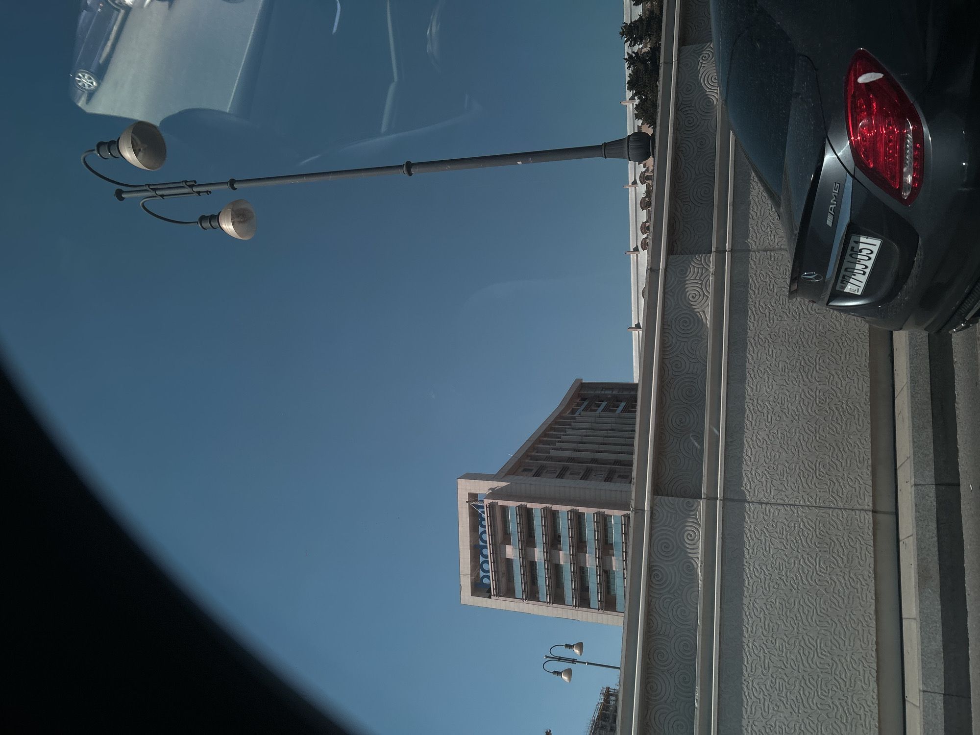 A dark gray Mercedes-Benz C-Class sedan with "AMG" badging is parked on the right side of the frame. The car's license plate reads "77-DJ-051". In the background, a modern, light-colored building with multiple floors and balconies is visible under a clear blue sky. A streetlamp with two lights stands to the left of the building. The scene is captured from a slightly elevated perspective, possibly from another vehicle, as indicated by the dark, curved edge framing the top left of the image. The architectural style of the building and the license plate suggest the location is Baku, Azerbaijan.